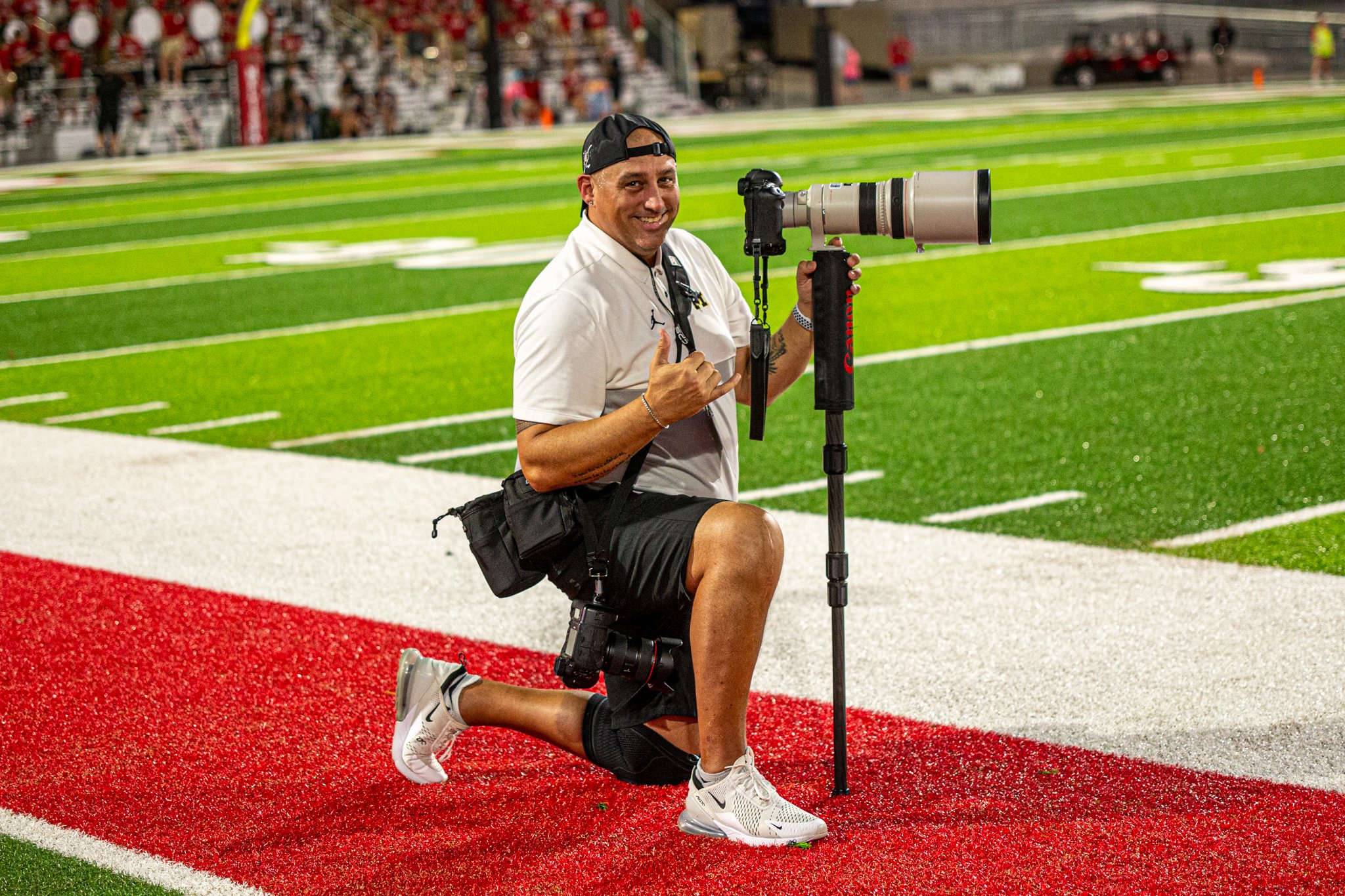 A man kneeling on a running track with a camera with a large telephoto lens, giving a thumbs-up and smiling, at a sports stadium with spectators in the background.