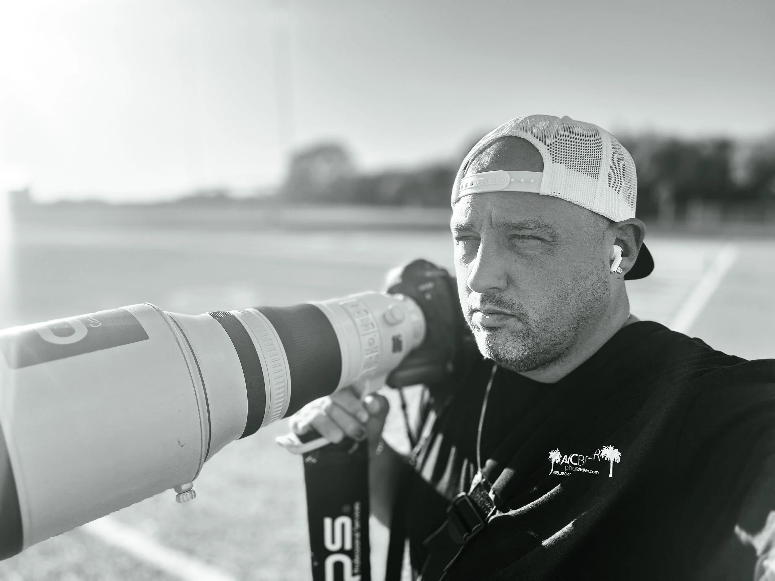 A person wearing a white trucker hat and a black jacket lensing a telescope outdoors in a bright setting with a blurred landscape in the background.