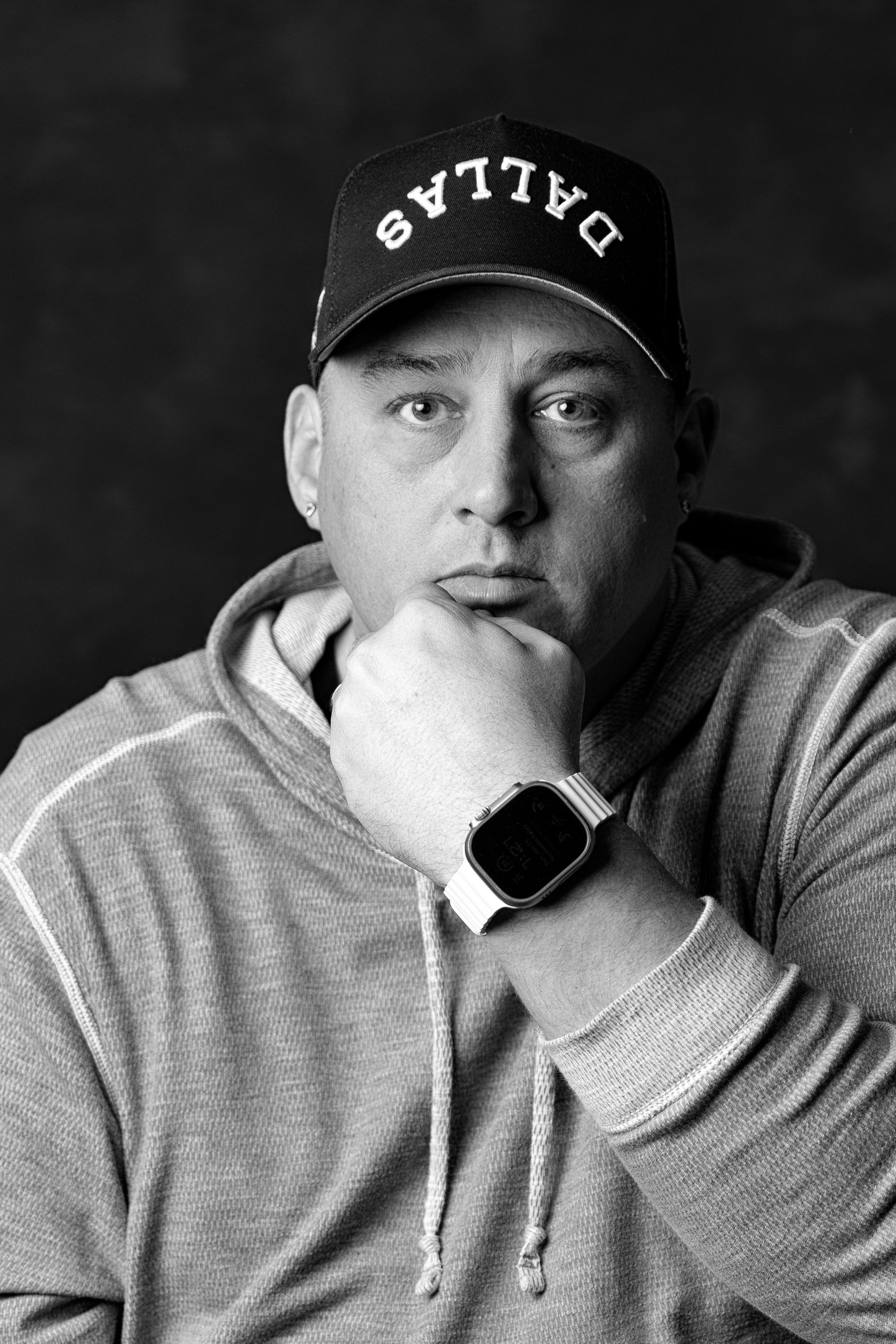 Black and white portrait of a person wearing a cap with the word 'DALLAS' on it, a hoodie, and a smartwatch on their wrist, with their hand resting on their face against a dark background.