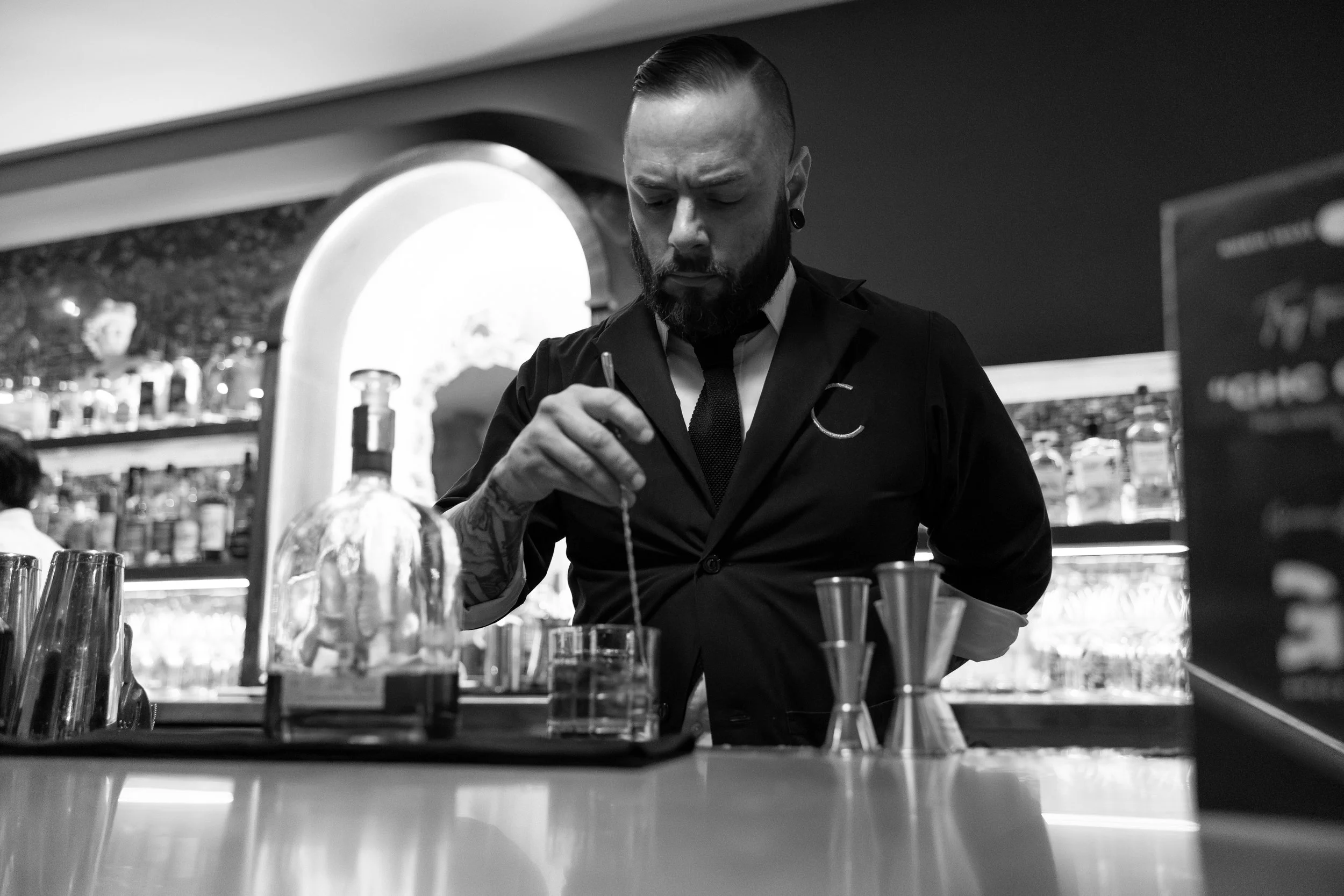 A man with a beard and piercings working behind the bar in a bar or restaurant, preparing drinks with bottles and mixing tools on the counter.