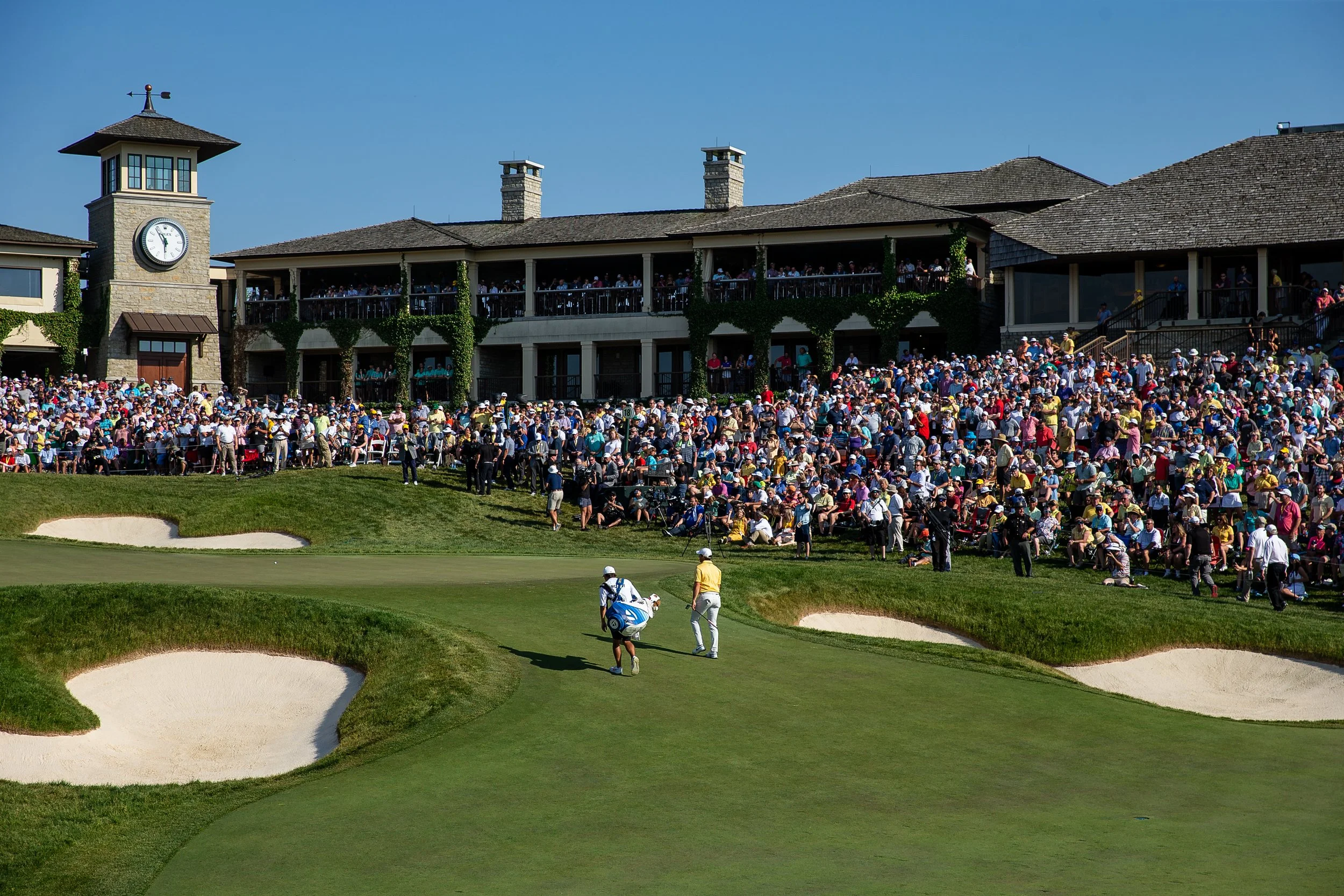 The image shows a large crowd of people gathered on a hillside, watching a golf tournament at a golf course with sand bunkers in the foreground. There is a building with a clock tower and a porch with people on it in the background under a clear blue