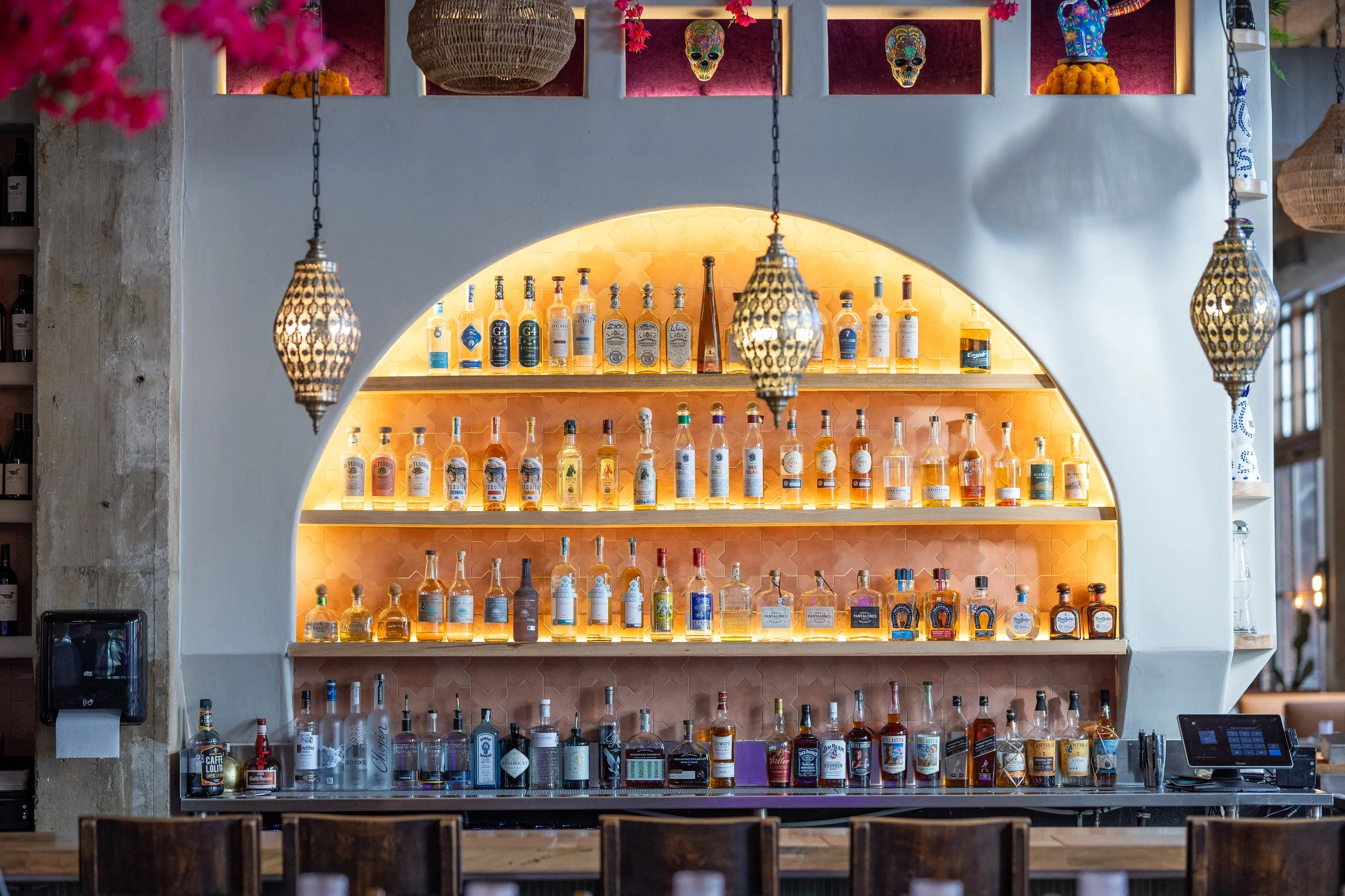 Bar with illuminated shelves stocked with various bottles of alcohol, decorative hanging lamps, and colorful skull artwork on the wall.