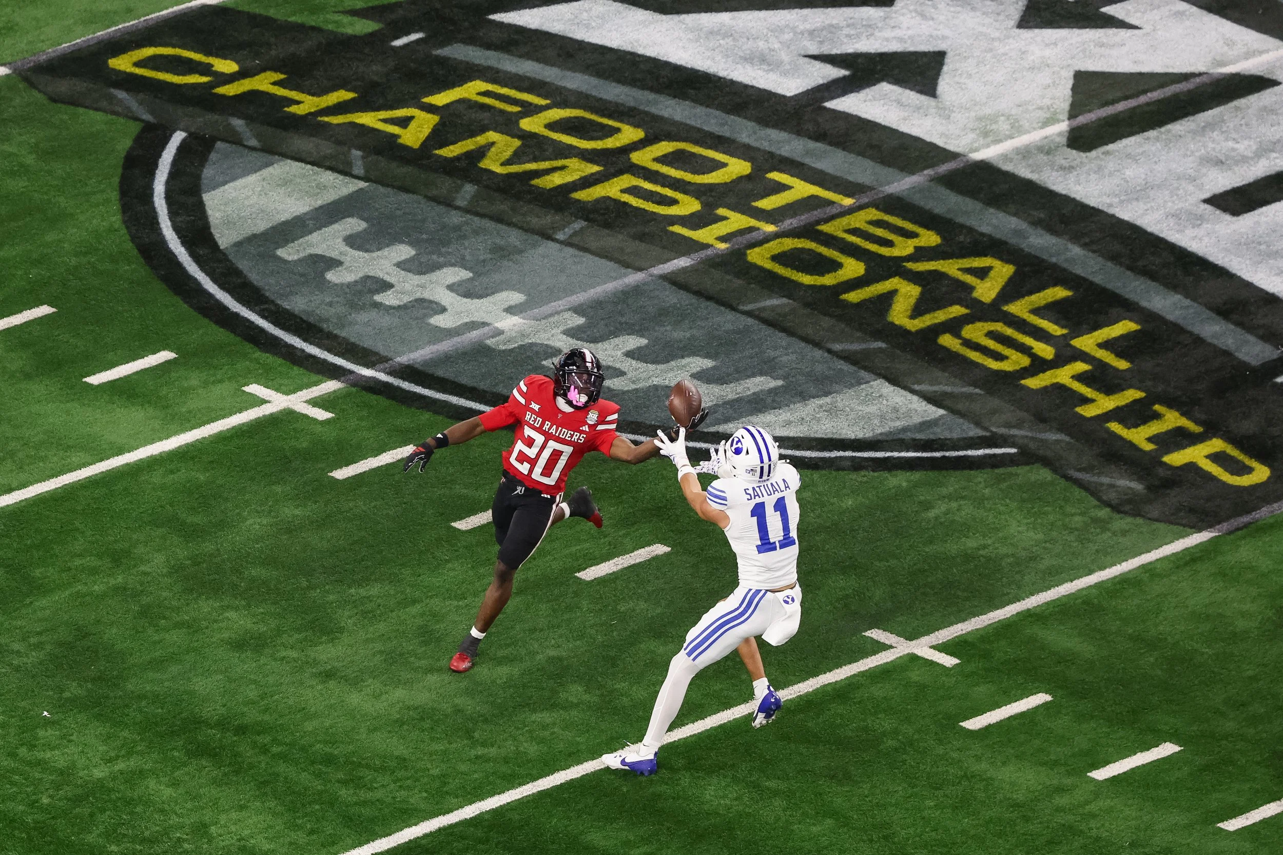 An American football game in progress with a player in a red jersey catching the football while a player in a white jersey defends, on a field with a large black and gray logo in the background.