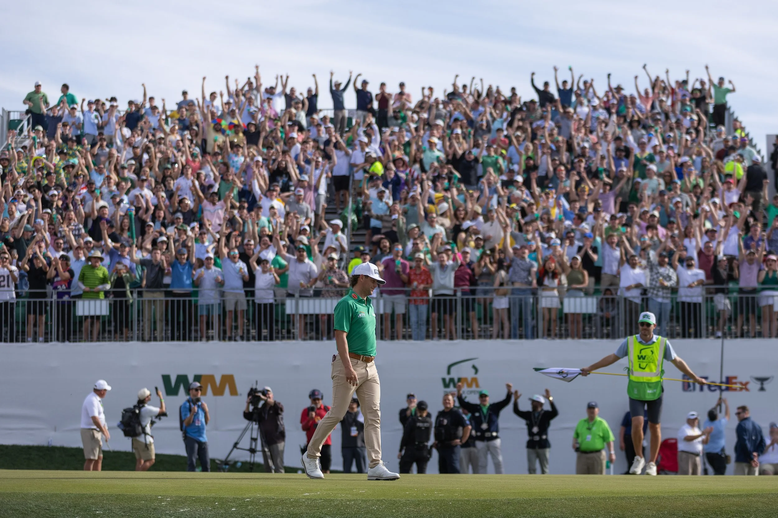 A golf course with a large crowd of spectators in the background and a golfer walking on the green in the foreground.