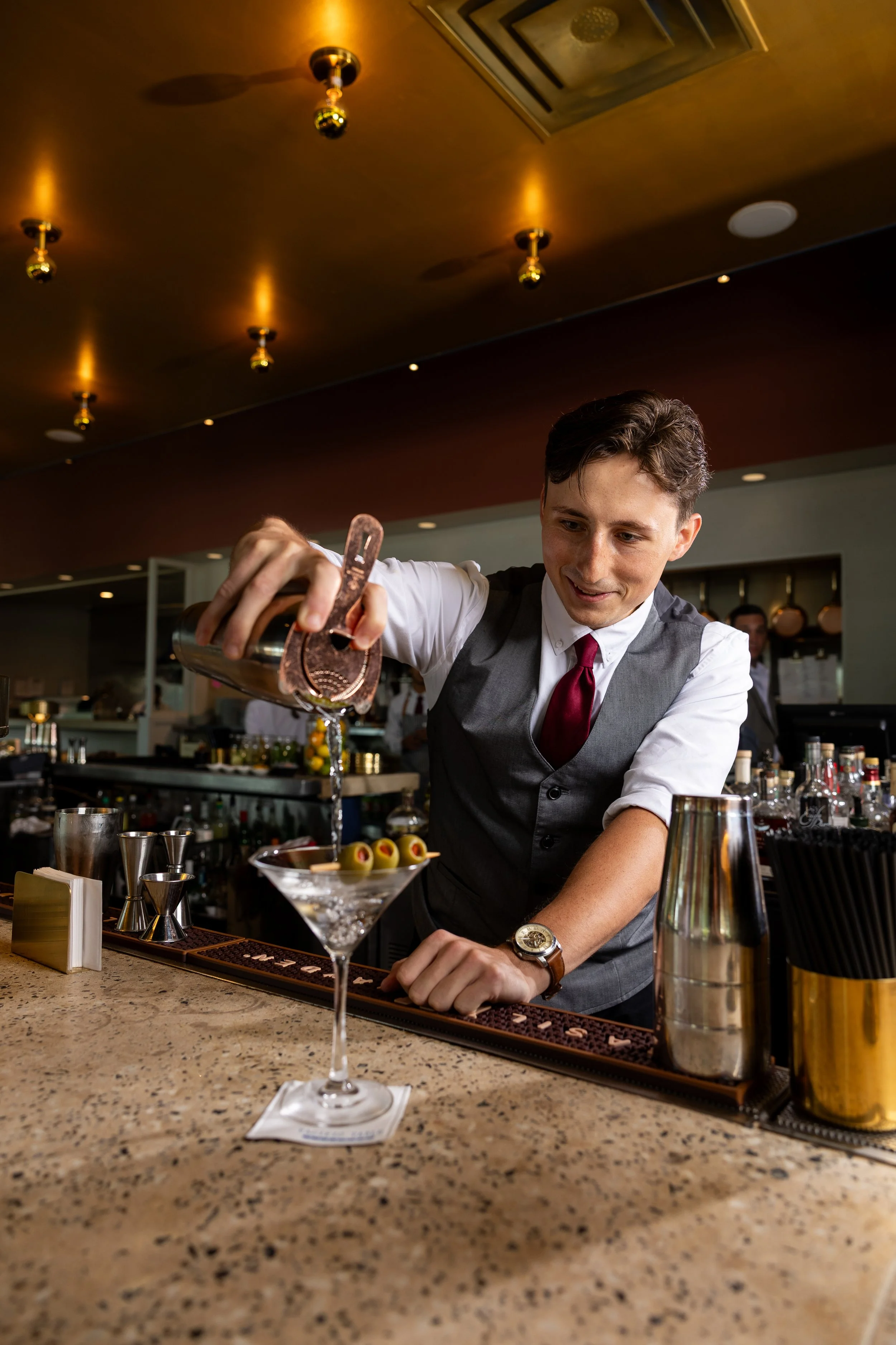A young male bartender preparing a cocktail at a bar counter, with various bar tools, drinks, and garnishes around him, in a dimly lit bar or restaurant setting.