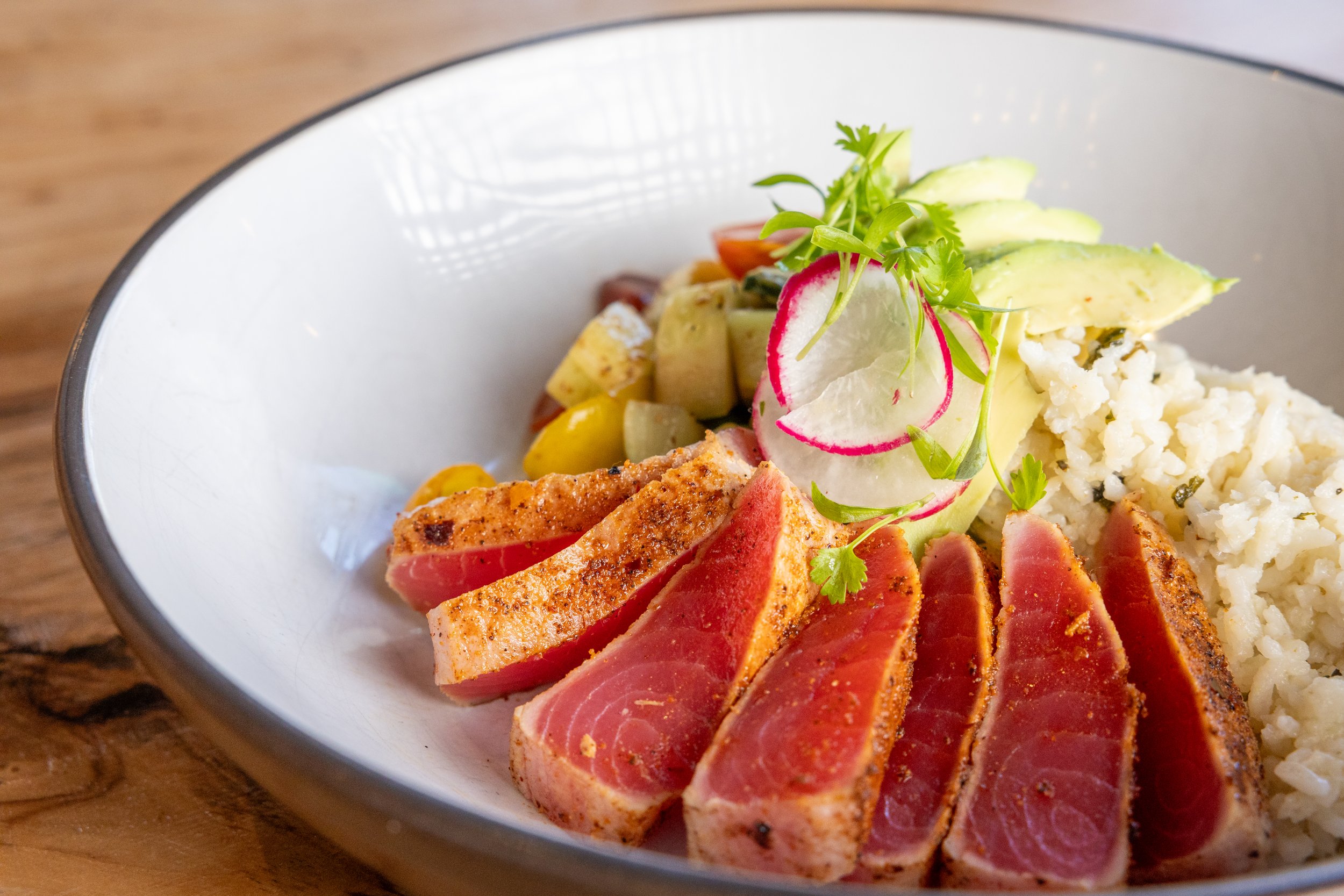 Sliced seared tuna with herbs, sliced radishes, avocado, mashed potatoes with herbs, cherry tomatoes, and microgreens in a white bowl on a wooden table.