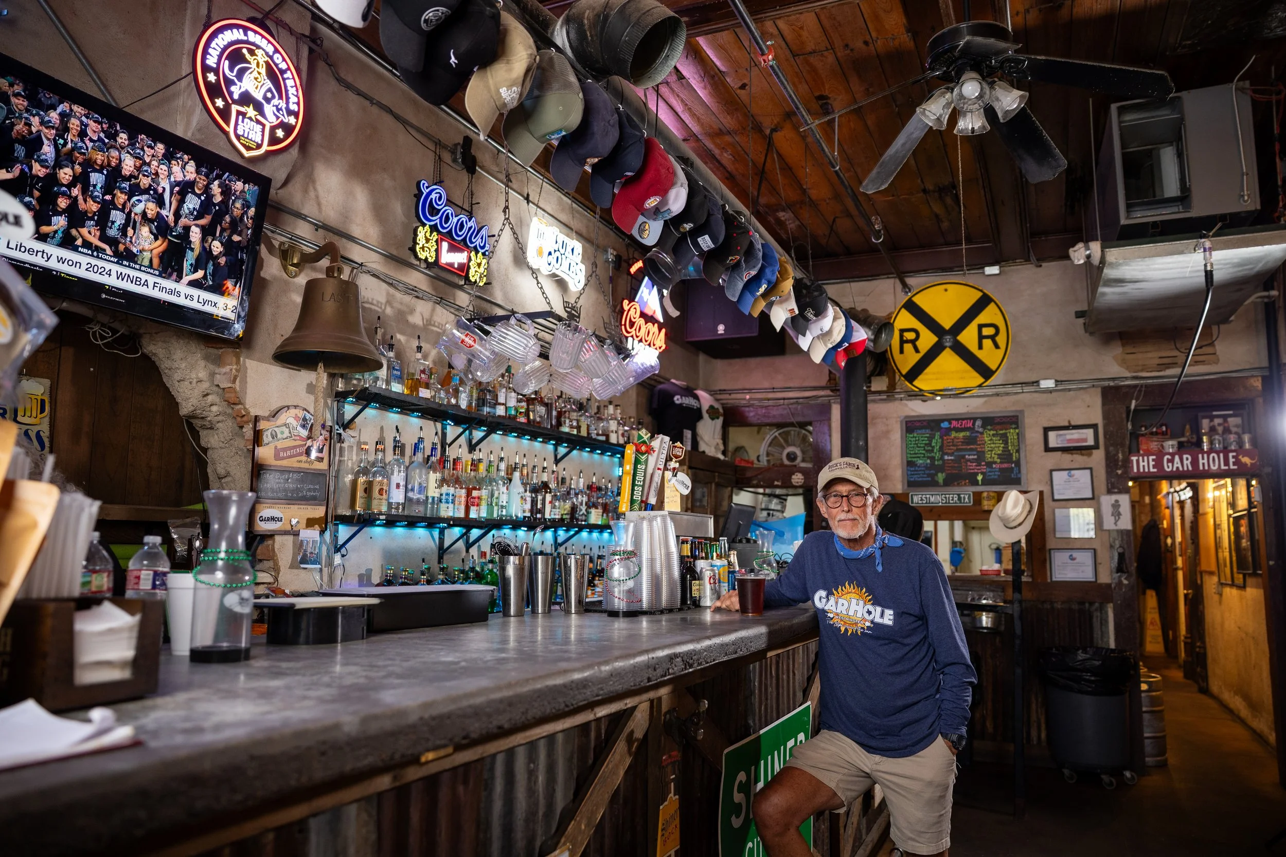 A man wearing glasses, a cap, and a blue Garhól T-shirt stands at a bar counter in a rustic tavern, with hats hanging from the ceiling and neon signs displayed behind the bar. The bar area is cluttered with bottles, glasses, and bar equipment, and a 