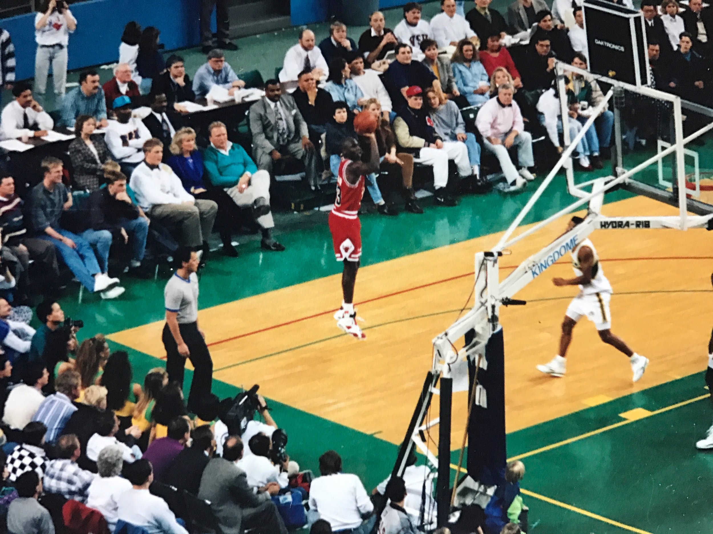 A basketball game with a player in a red uniform jumping to shoot the ball, and a crowd of spectators watching from the stands.