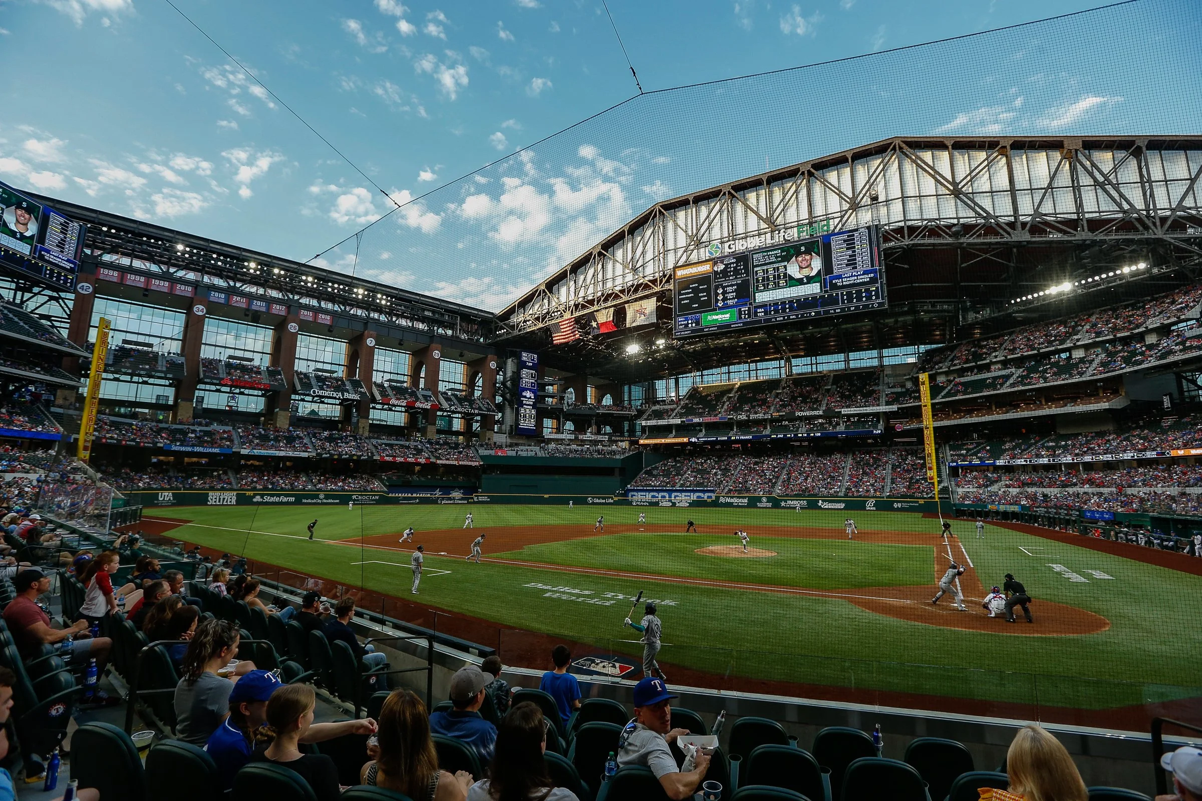 A baseball game is taking place at Globe Life Field stadium. Spectators are seated in the stands, watching the game, while players are on the field. The stadium has a large digital scoreboard displaying game information, and the sky is blue with some