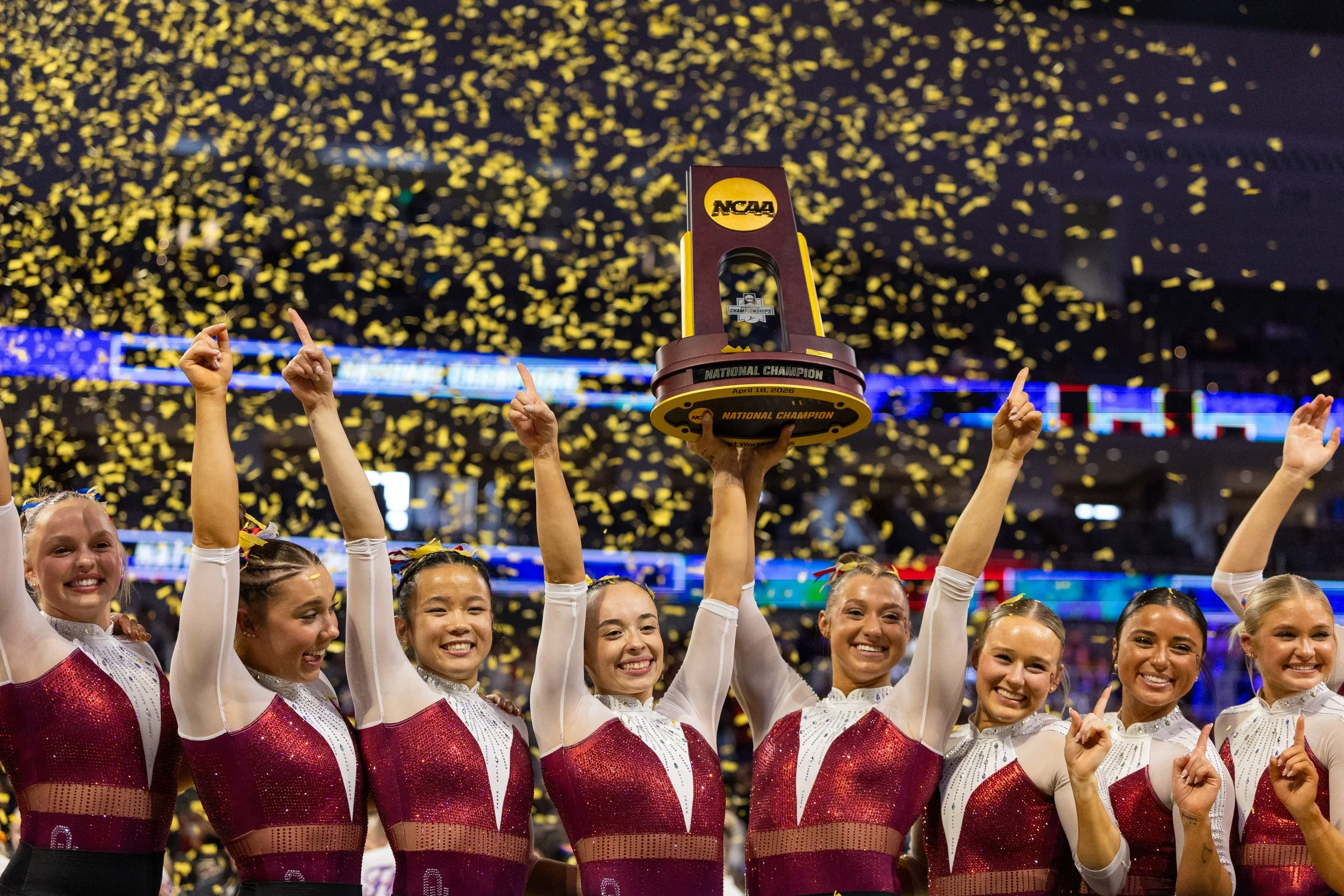 A group of female gymnasts in maroon and white leotards celebrating on a gymnastics competition floor, holding up a trophy with yellow confetti falling in the background.