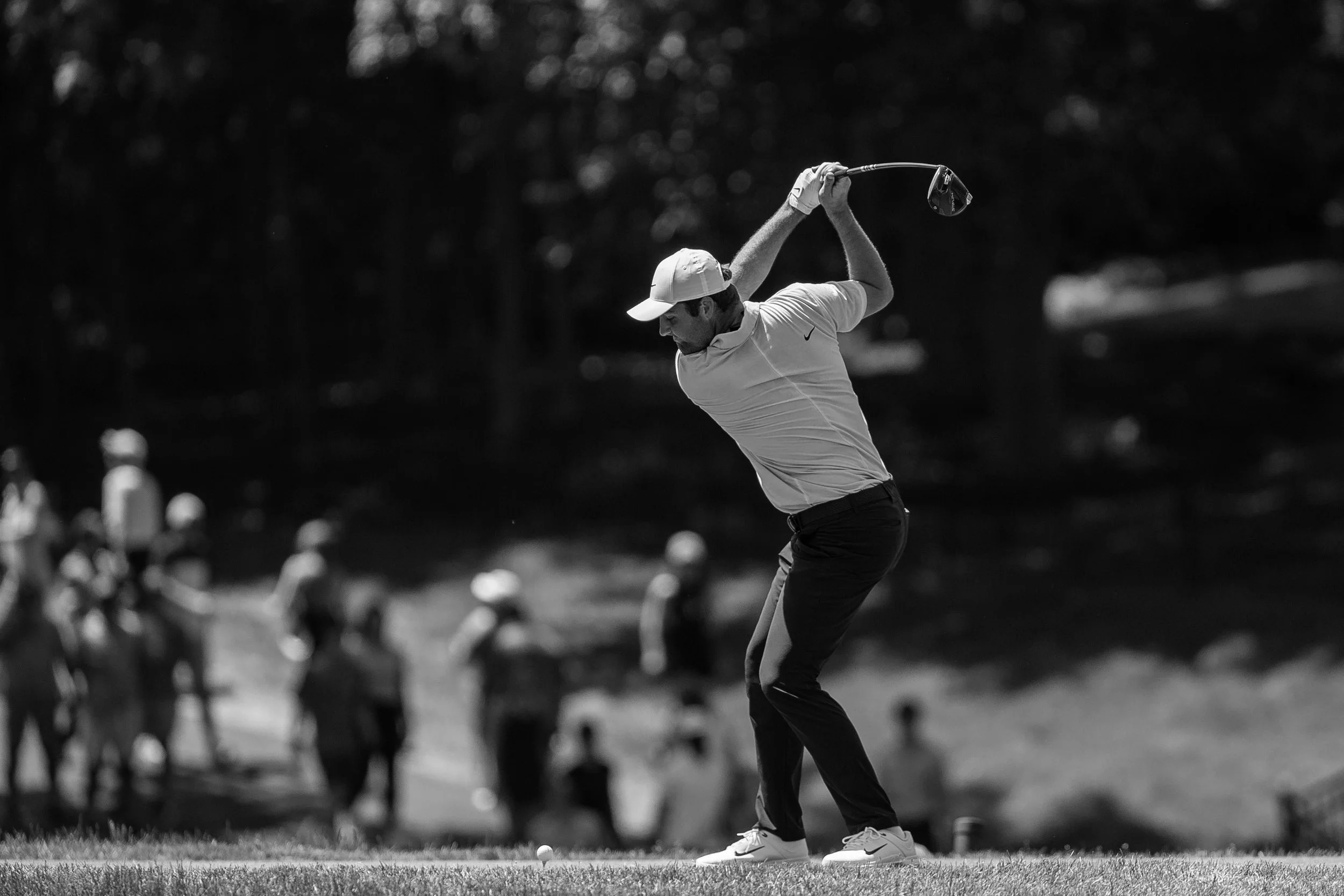 A man in a white Nike shirt and cap playing golf, swinging a club on a golf course with a golf ball on the ground.
