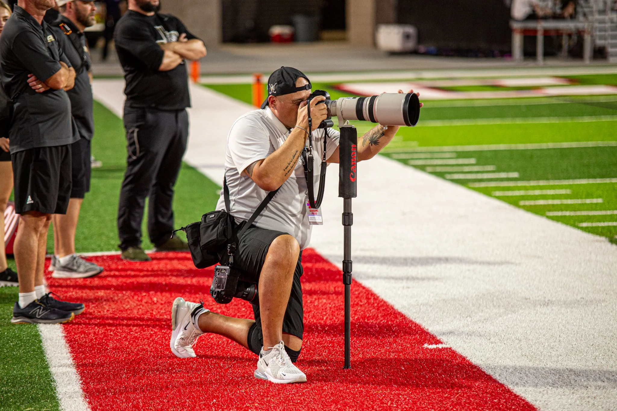 A photographer kneeling on one knee on a football field, taking pictures with a large telephoto camera lens mounted on a tripod, while other people stand nearby watching. The field has green artificial turf with red and white yard lines.