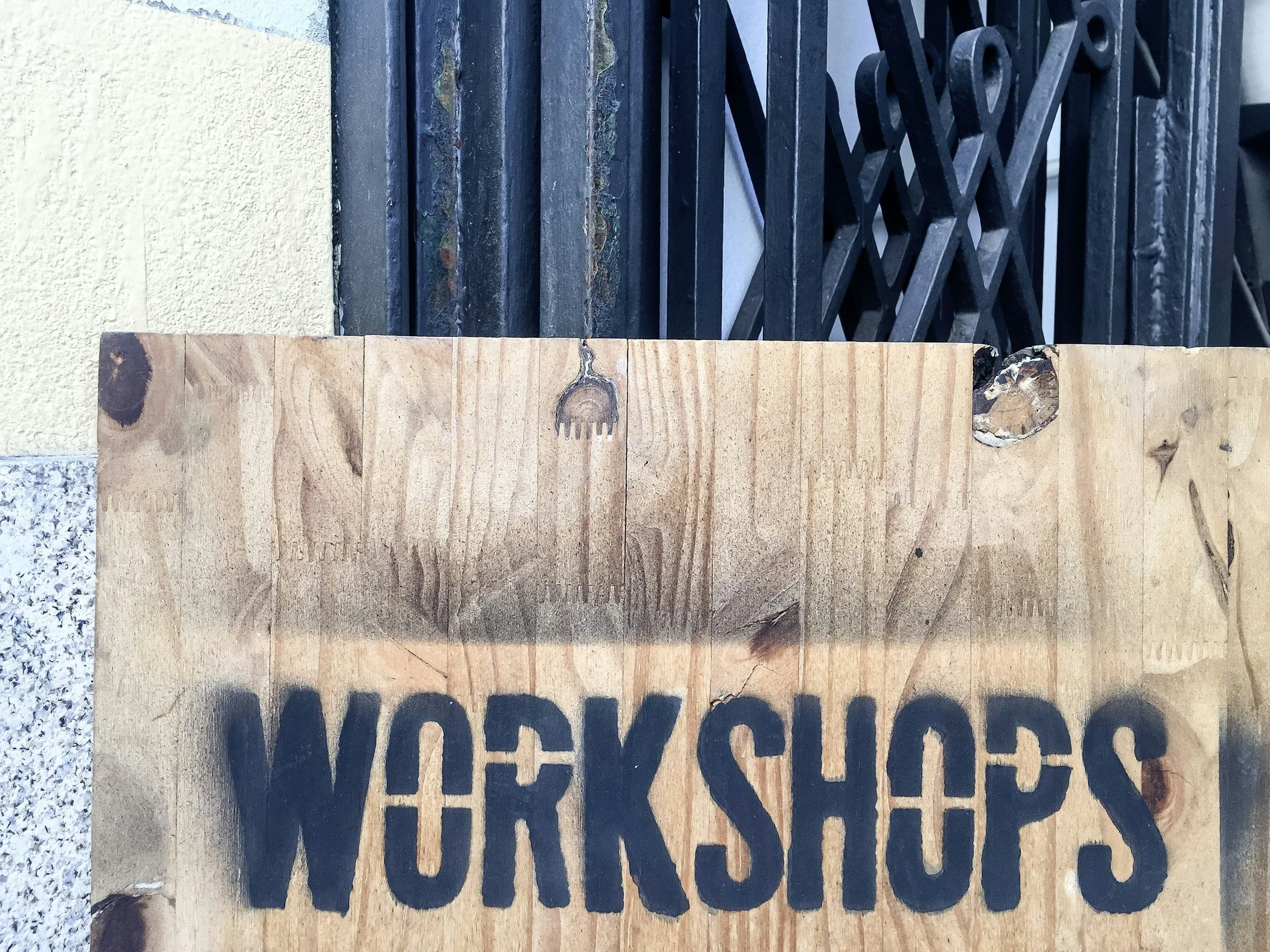 A wooden board placed upright with black stencil letters spelling 'WORK' on it, with an iron gate and wall in the background.