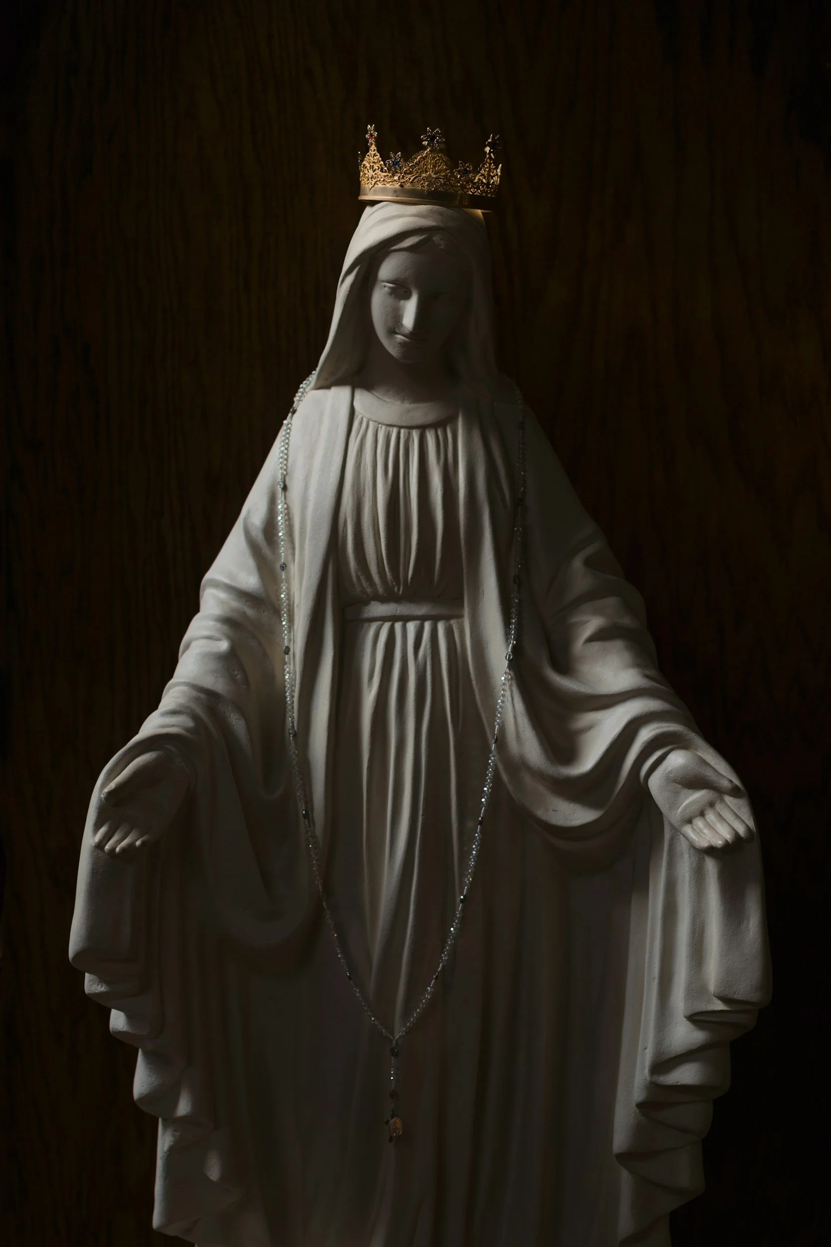Statue of the Virgin Mary with a golden crown, draped in white, with a dark background.