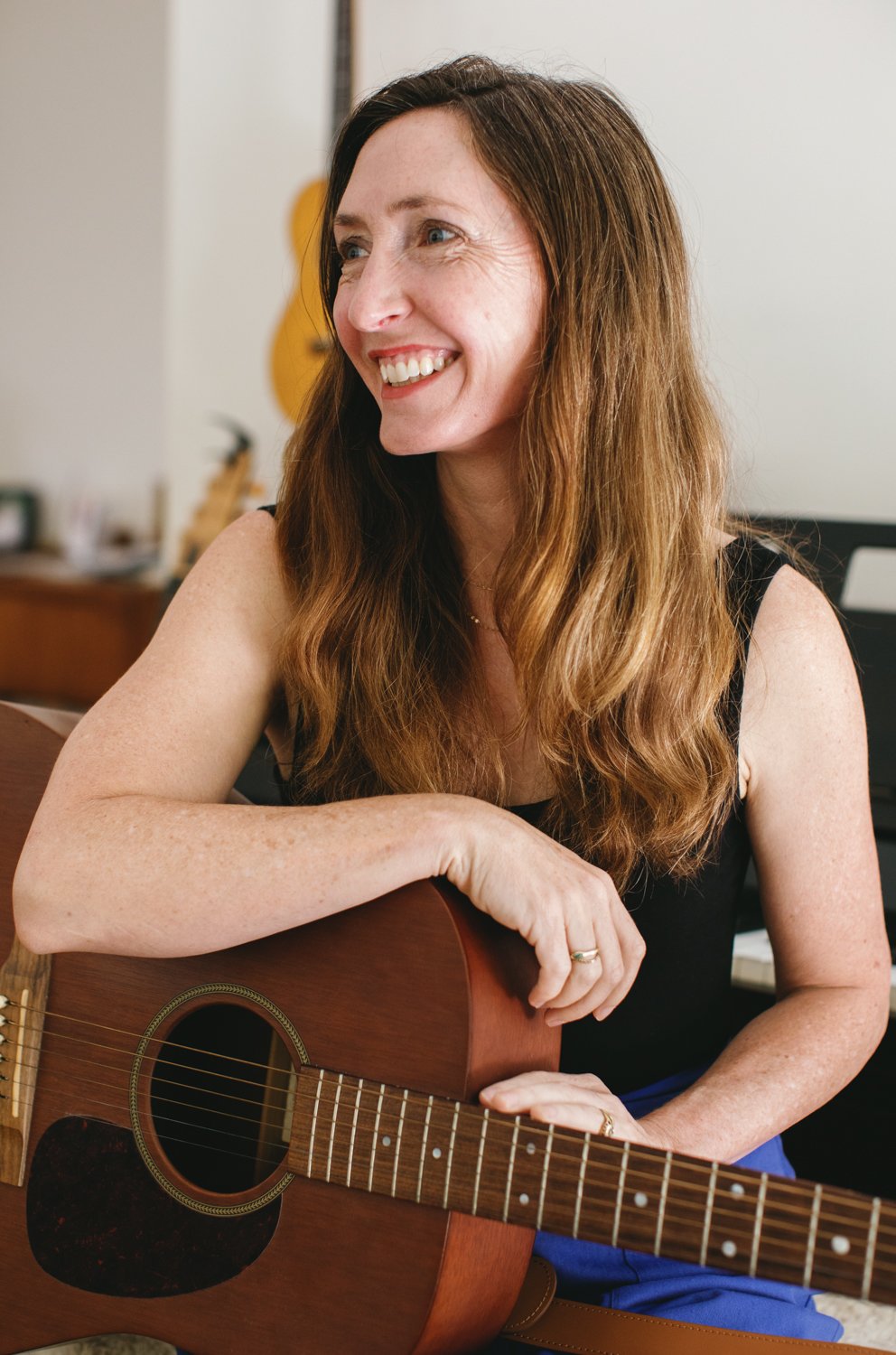 A woman with long red hair smiling and sitting with an acoustic guitar in a room.