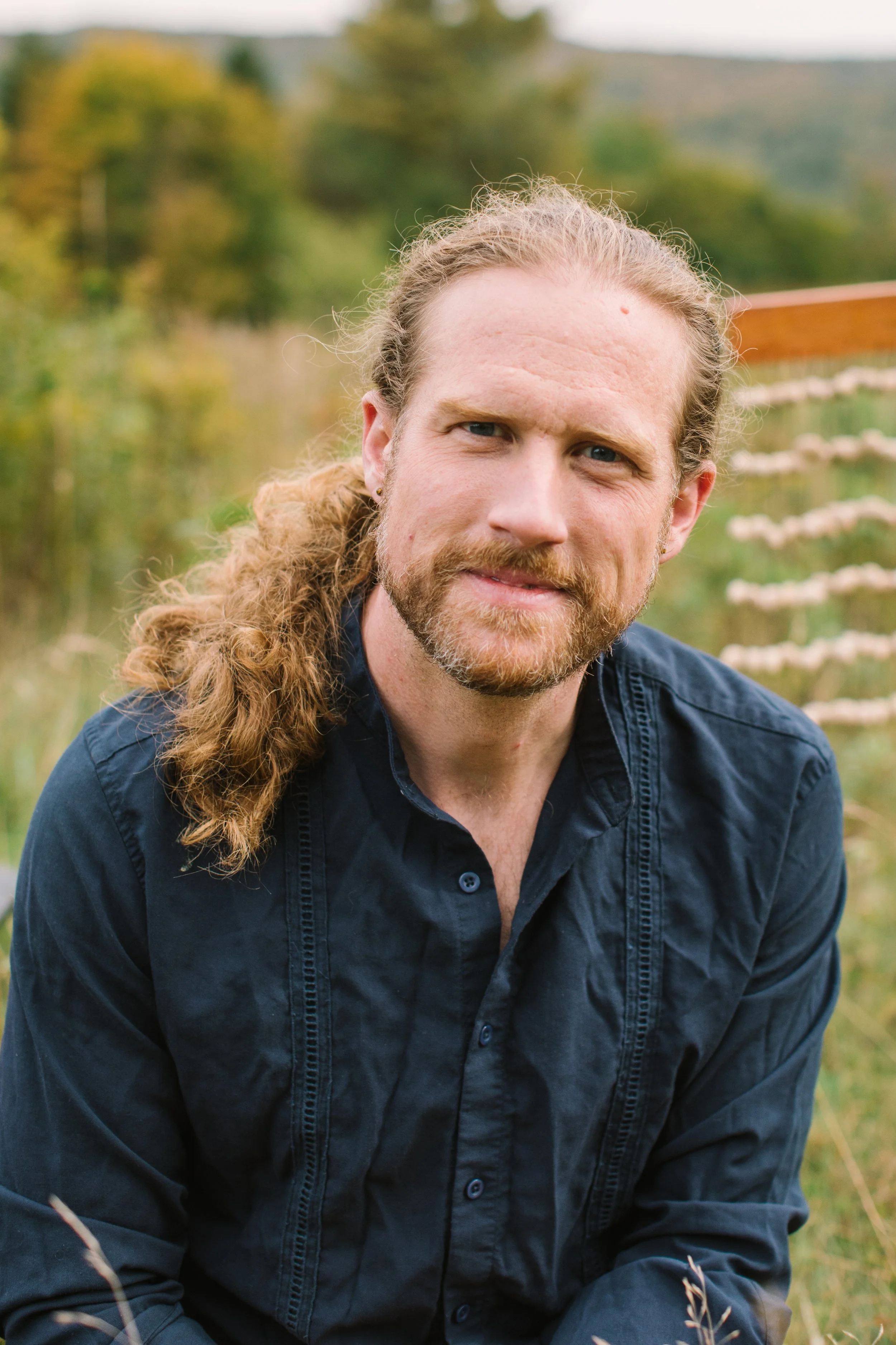 A man with long curly hair, a beard, and light skin, wearing a dark blue button-up shirt, is outdoors in a park with green trees and a wooden fence in the background.