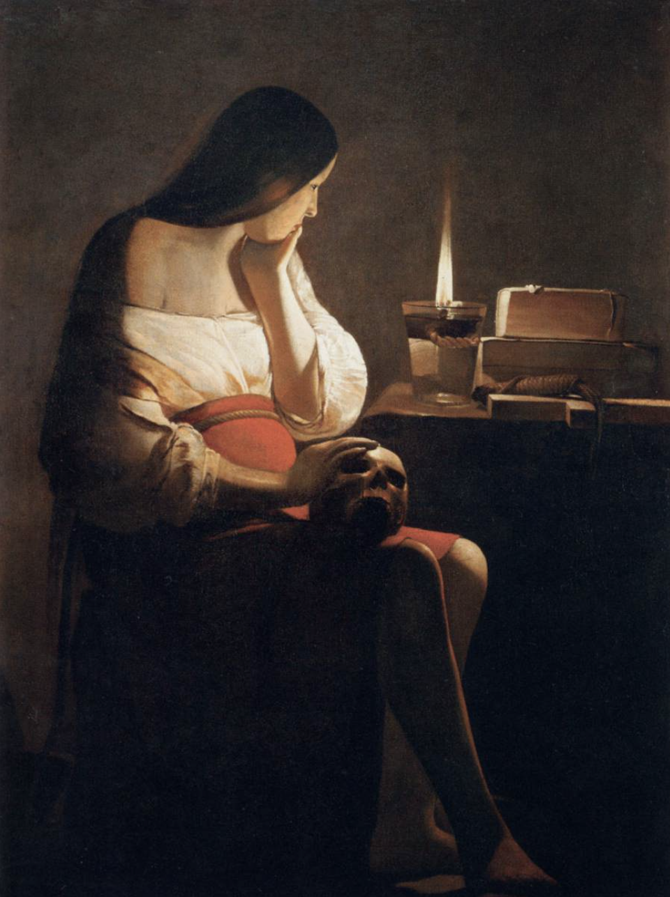 A woman sitting in a dark room holding a human skull while looking at an open book and a lit candle on a table.