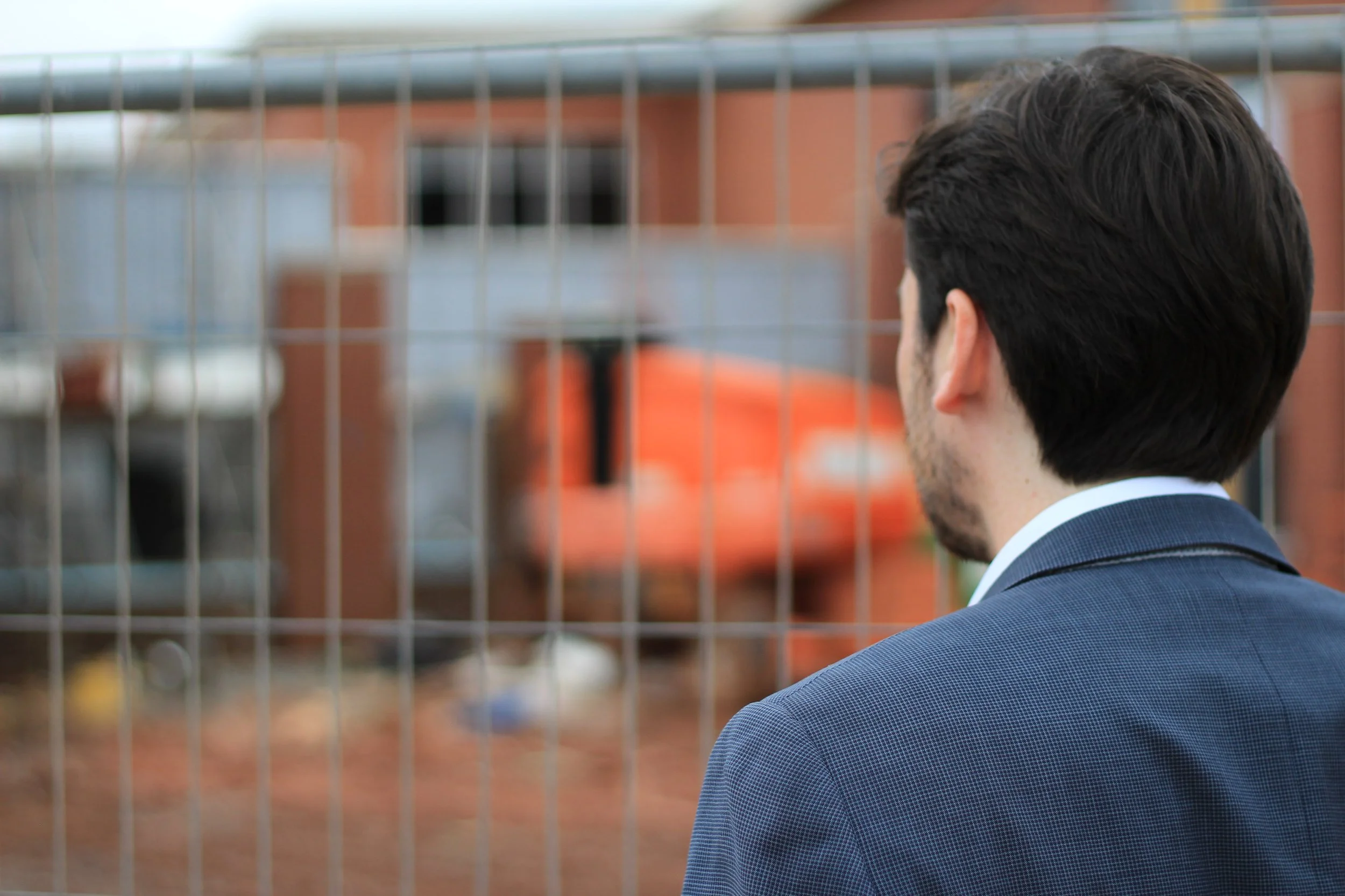 A man in a suit with dark hair stands behind a metal fence, looking toward a brick building with orange accents.