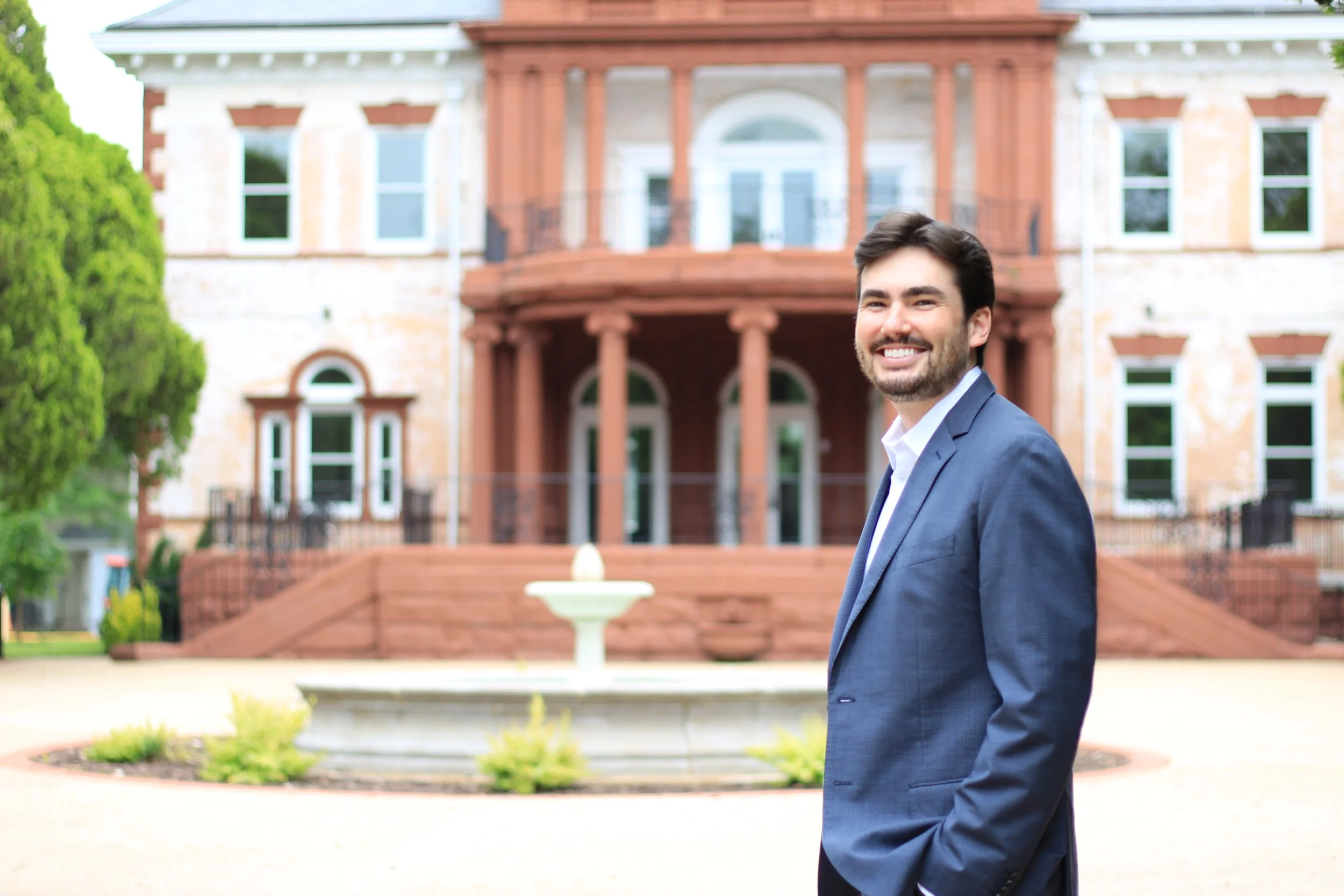 A man in a blue suit smiling outdoors in front of a historic brick mansion with a fountain and lush greenery.