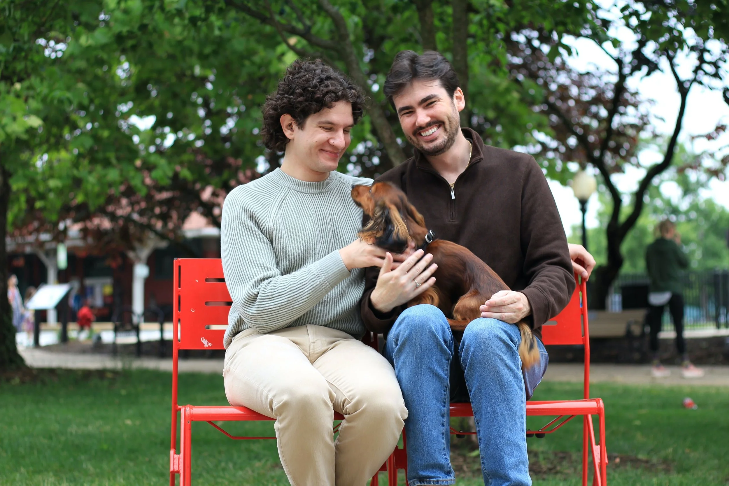 Two men sitting on a red bench in a park, holding a small dachshund dog, smiling and enjoying each other's company surrounded by green trees.