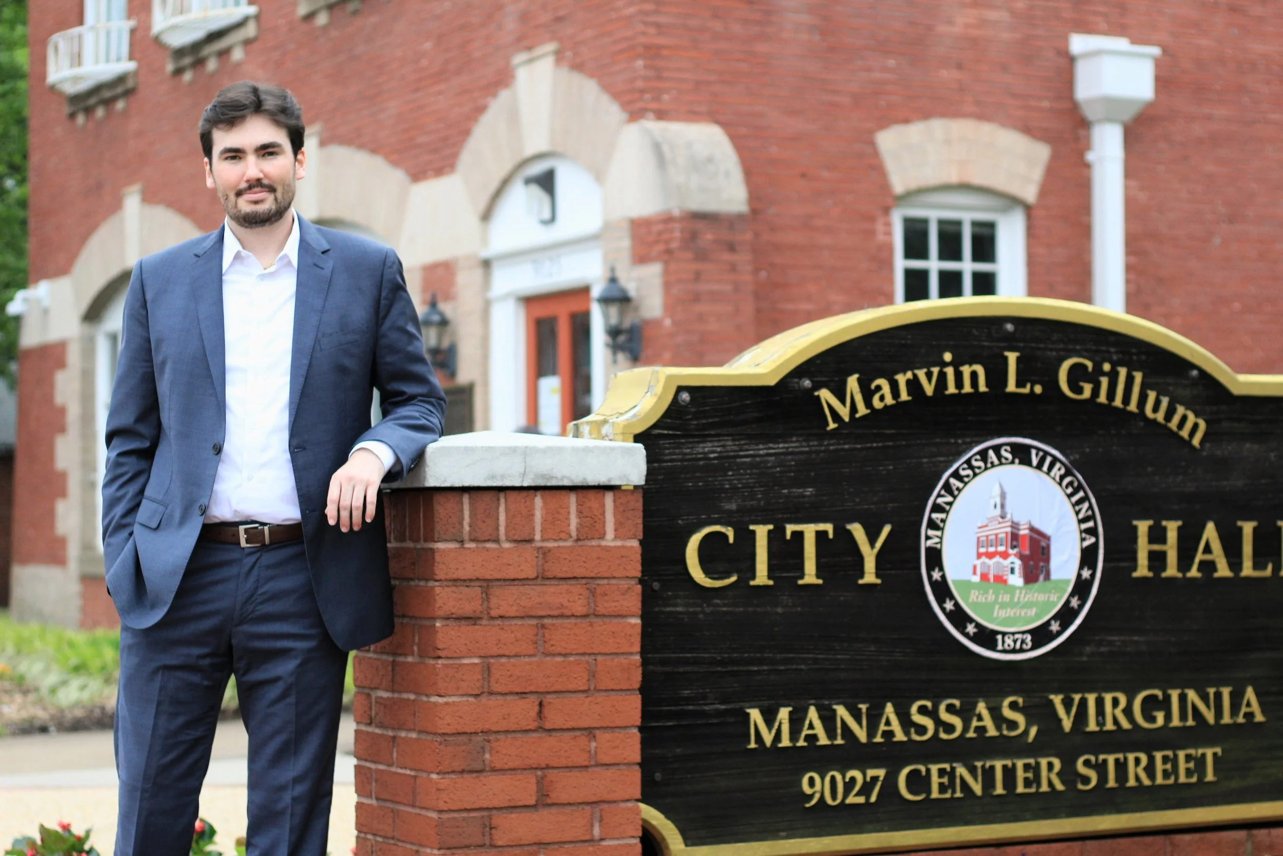 A man in a blue suit standing next to a brick sign on a sidewalk in front of a brick building with white trim and windows in Manassas, Virginia.