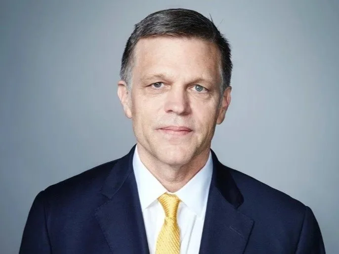 A middle-aged man wearing a dark suit, white shirt, and yellow tie, posing against a gray background.