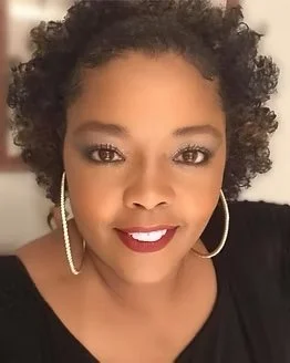 Close-up of a woman with curly hair wearing hoop earrings and a black top, smiling at the camera.
