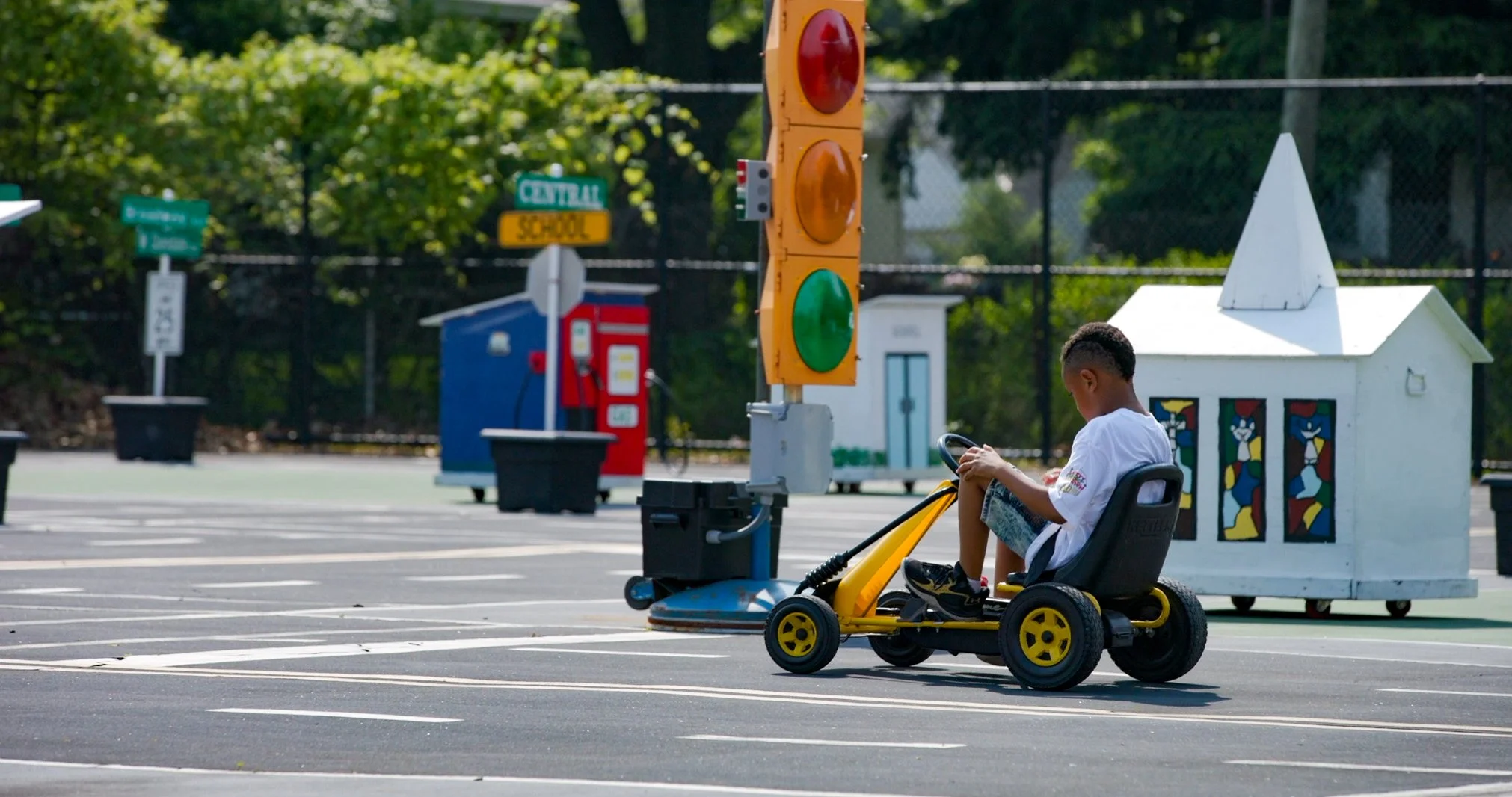 A young boy in Safety Town, sitting in a pedal car. In the background, there is a traffic light, a colorful small building with windows, and greenery with trees.