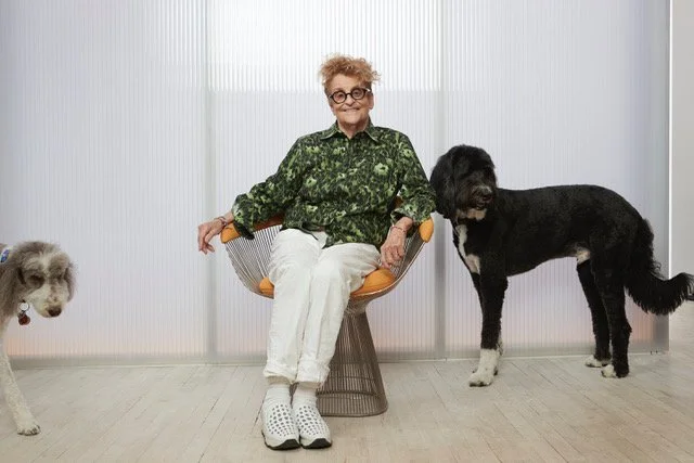 An elderly woman with curly hair and glasses sitting on an orange chair with wooden arms, wearing a green patterned shirt and white pants, smiling. There are two dogs, one large black and white dog standing next to her and a small light-colored dog partially visible on the left side of the image.