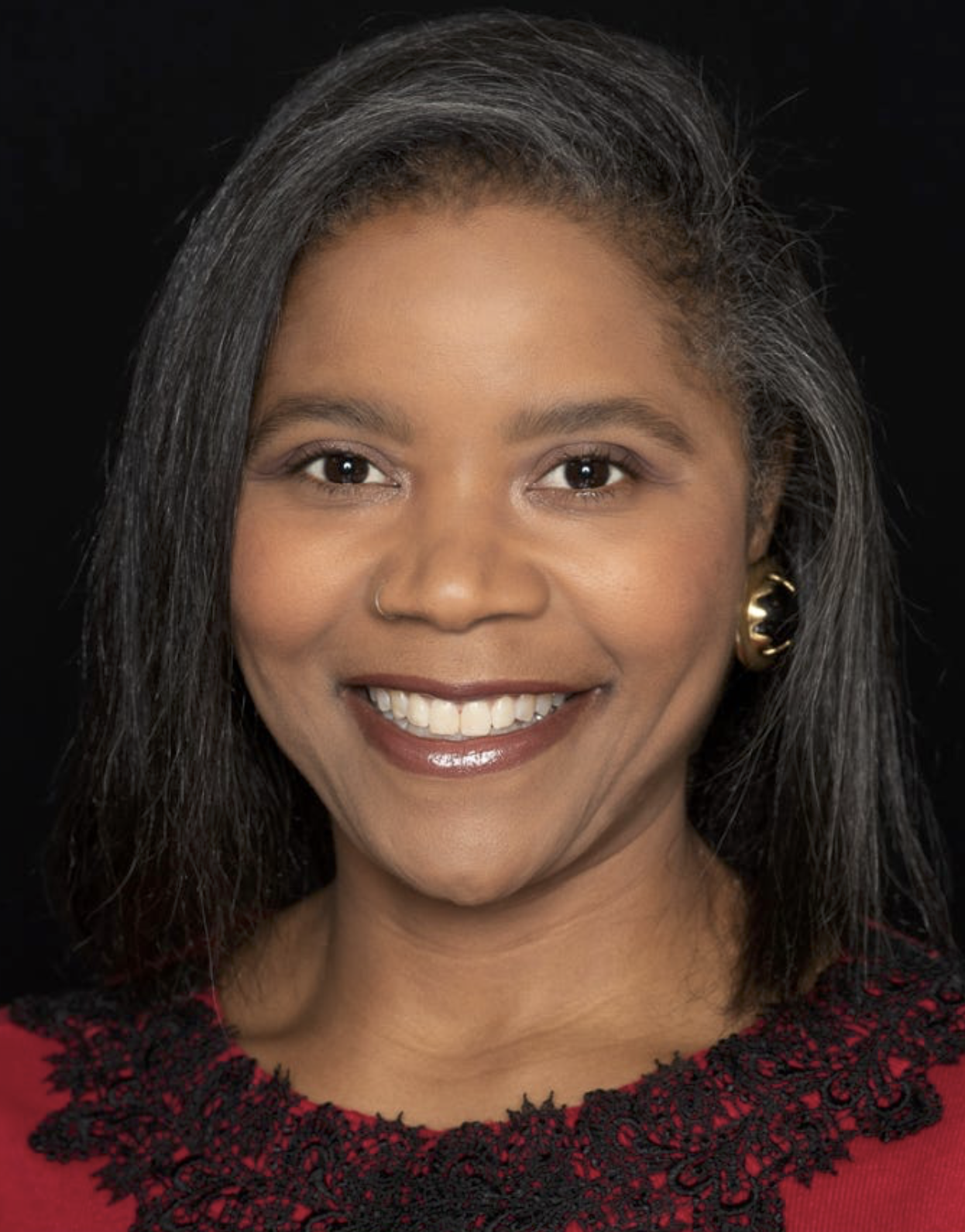 Close-up portrait of a woman with medium skin tone, long dark hair, gold earrings, smiling, wearing a red top with black lace details, against a black background.