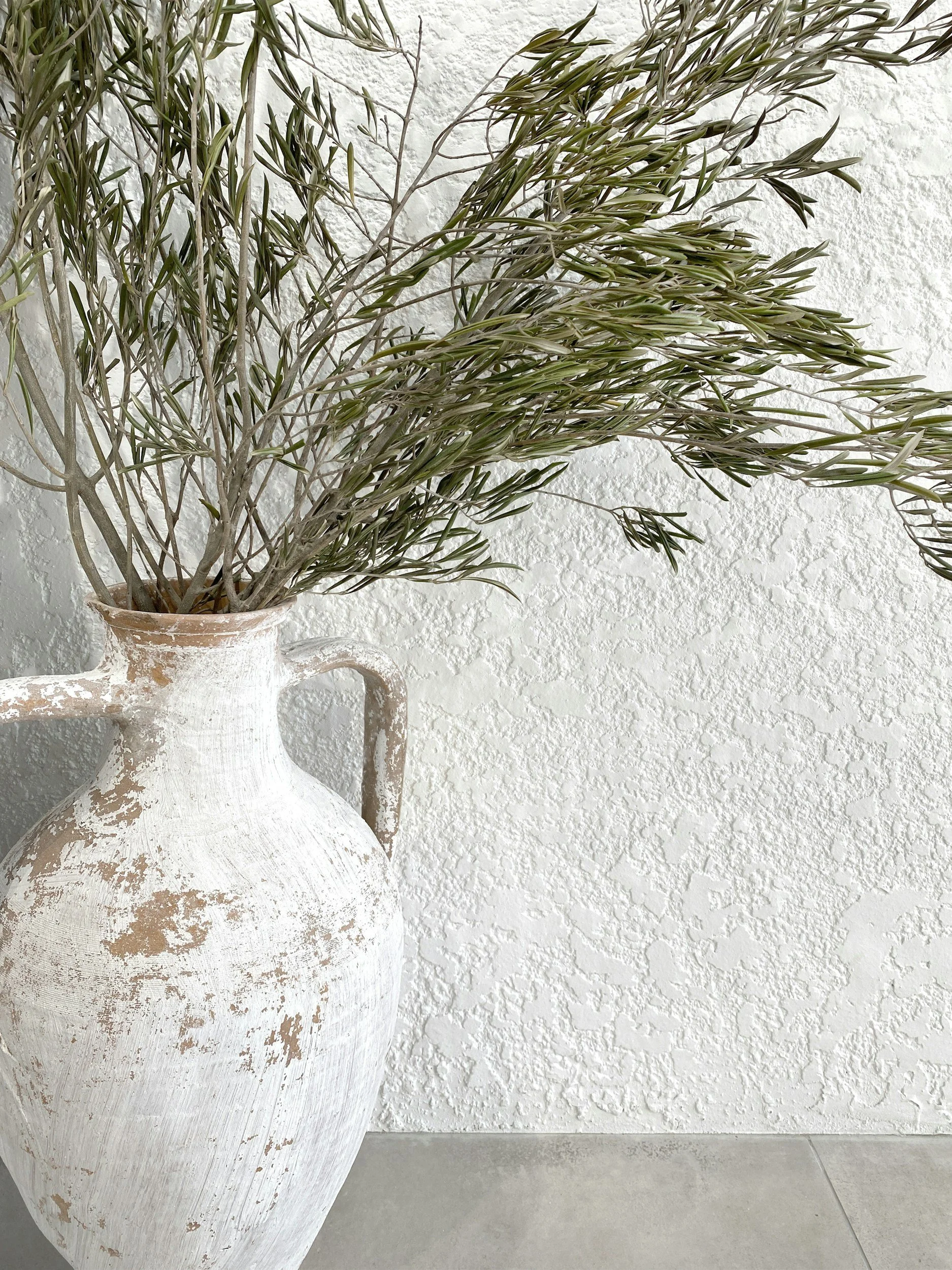 A large dried olive branch in a white, textured ceramic vase against a white textured wall.