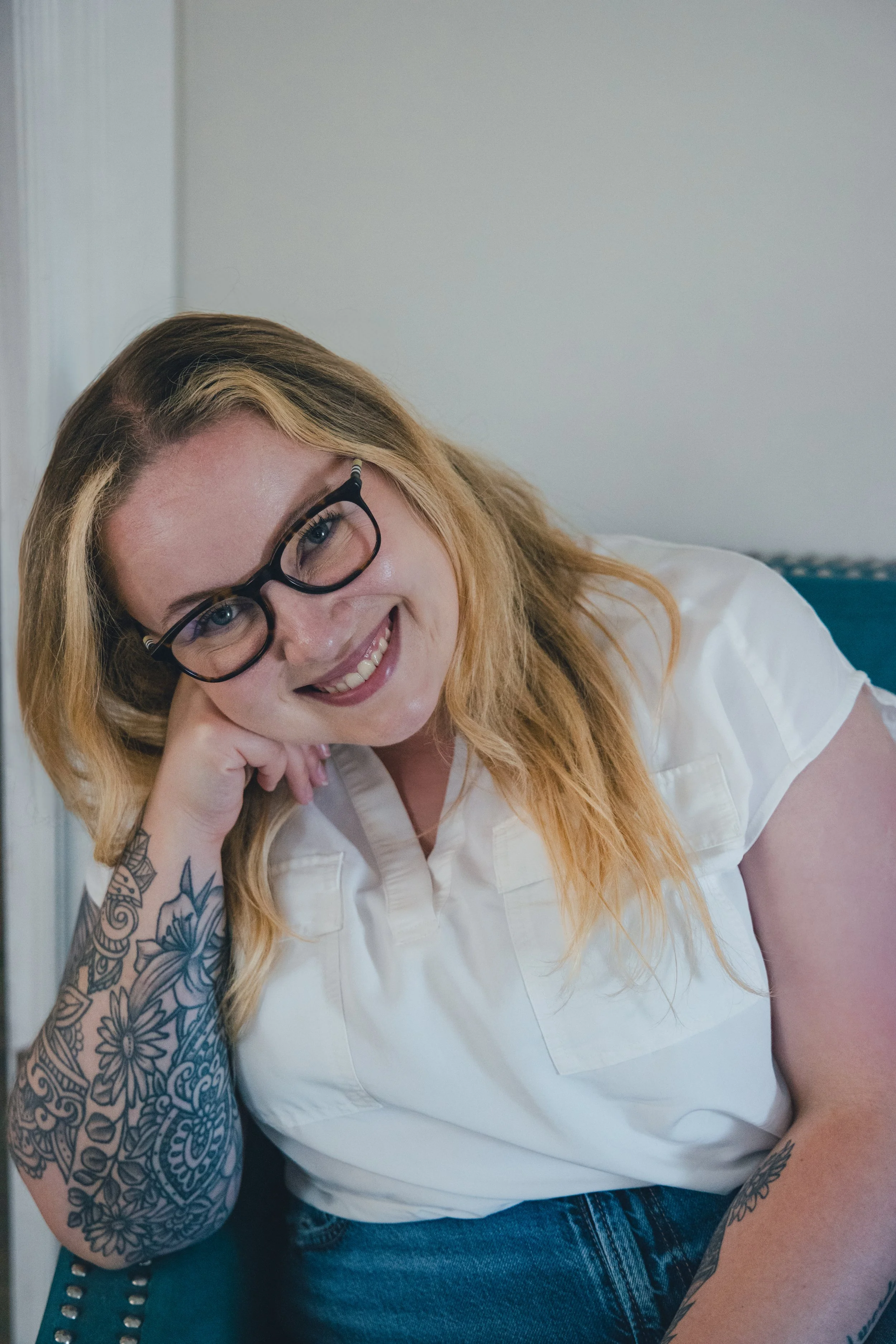 A woman with glasses and tattoos is sitting and smiling, wearing a white shirt and jeans.