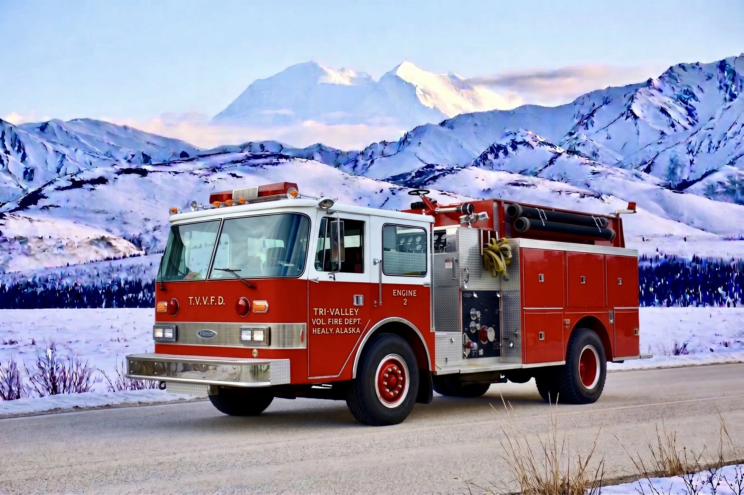 A red fire truck labeled 'Tri-Valley Volunteer Fire Department, Healy, Alaska' driving on a snowy road with snow-covered mountains and a partly cloudy sky in the background.
