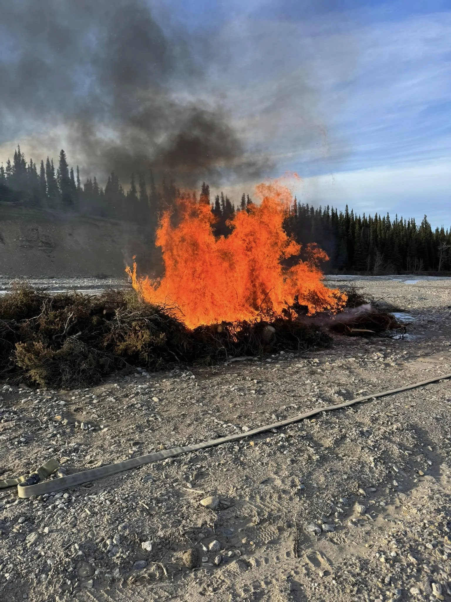 A wildfire burning in a forested area near a river, with thick black smoke rising into the sky. The ground is rocky and dry, with a hose lying on the terrain.