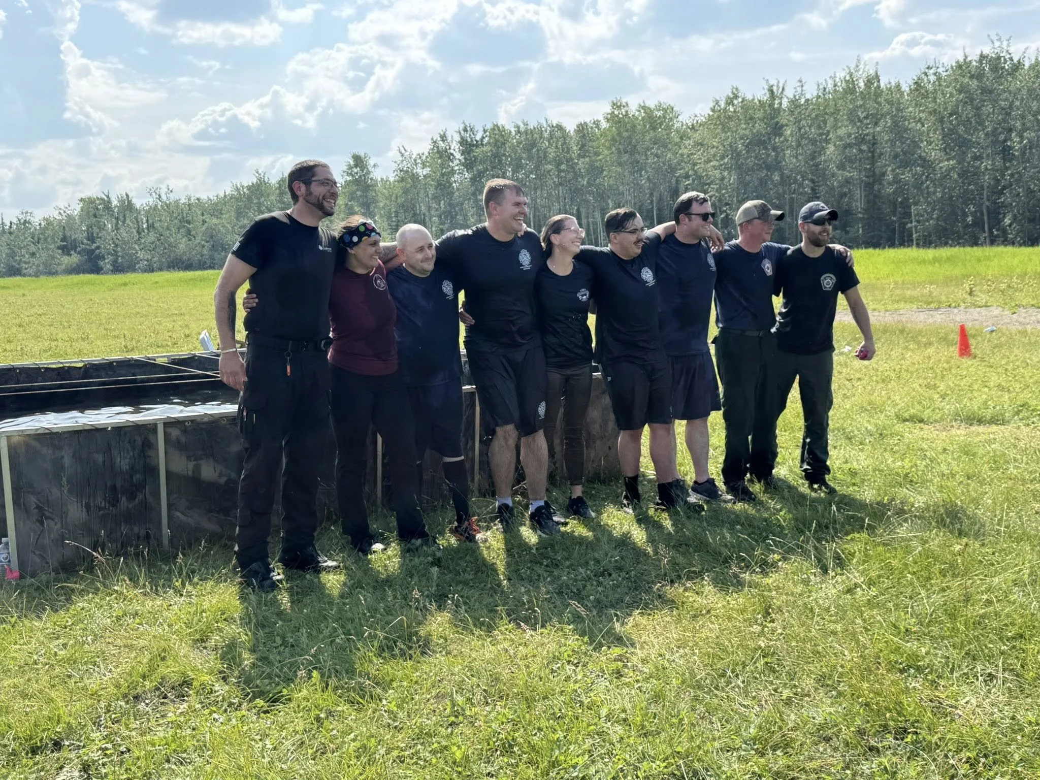 Group of people standing together outdoors in a grassy field with trees and blue sky in the background, smiling and embracing.