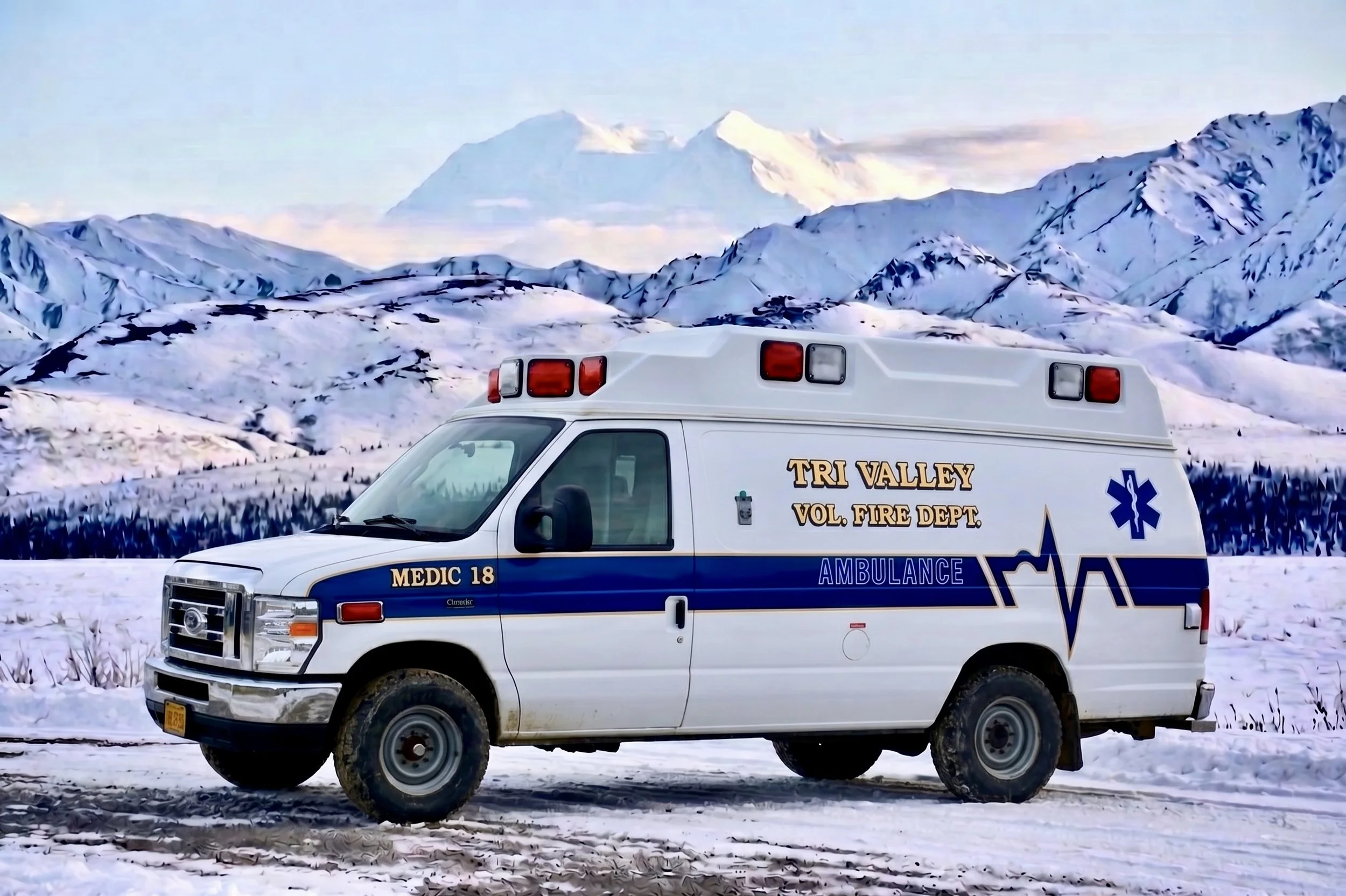 A white ambulance truck with blue and yellow markings, labeled 'Tri Valley Vol. Fire Dept.', parked on snow-covered ground with snow-capped mountains and a large mountain in the background.