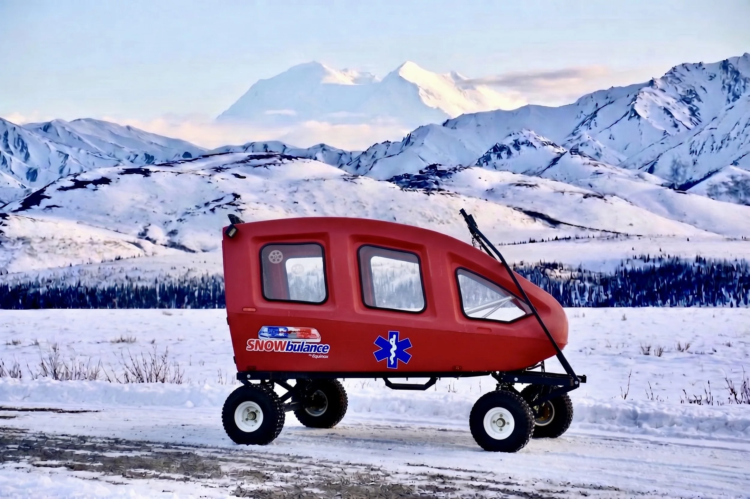 A red snow ambulance vehicle labeled 'SNOWbulance' with wheels, situated on snow-covered ground in a mountainous winter landscape with snow-covered peaks in the background.
