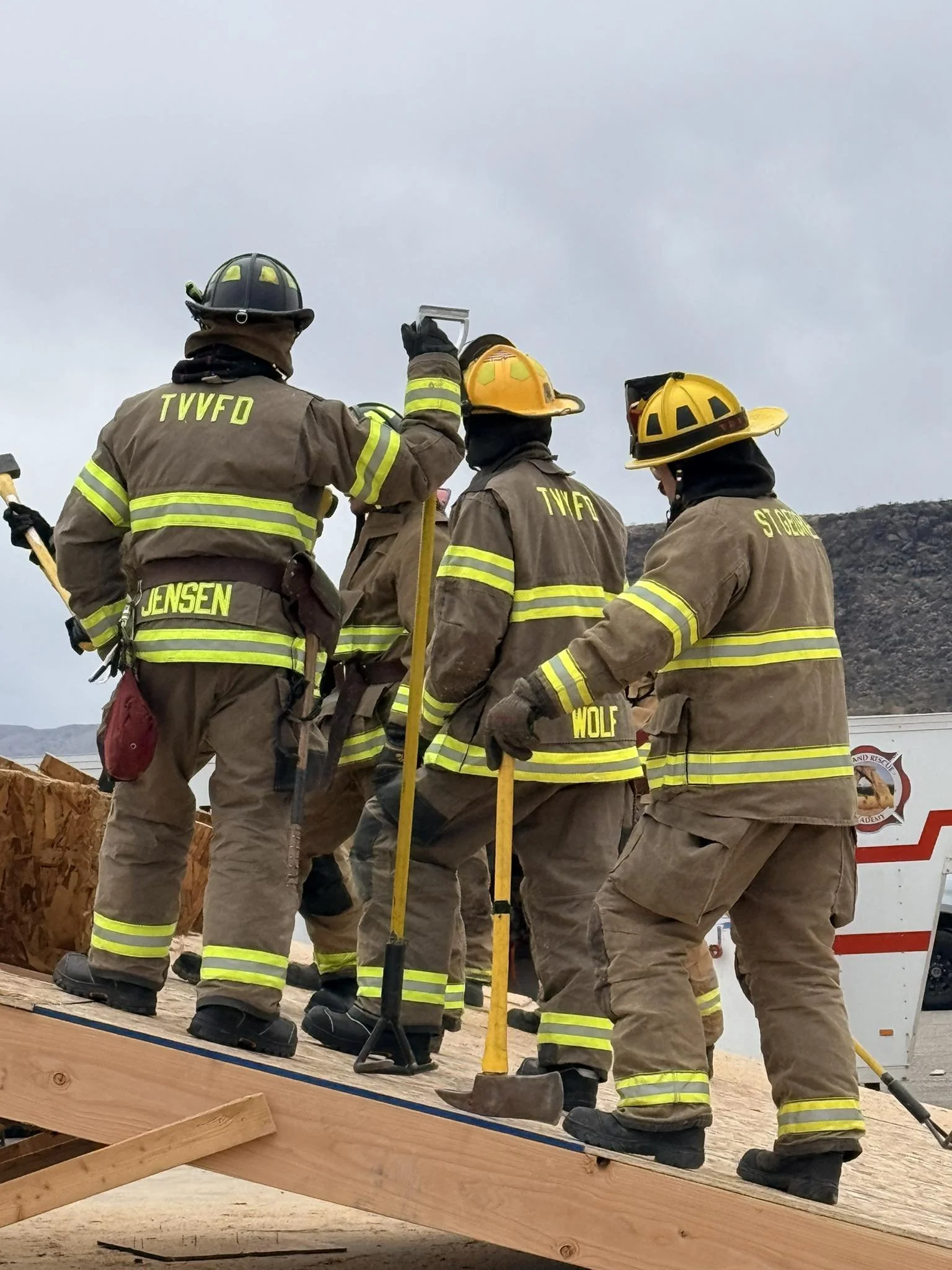 Group of firefighters in protective gear working together on a wooden structure outdoors during daytime.