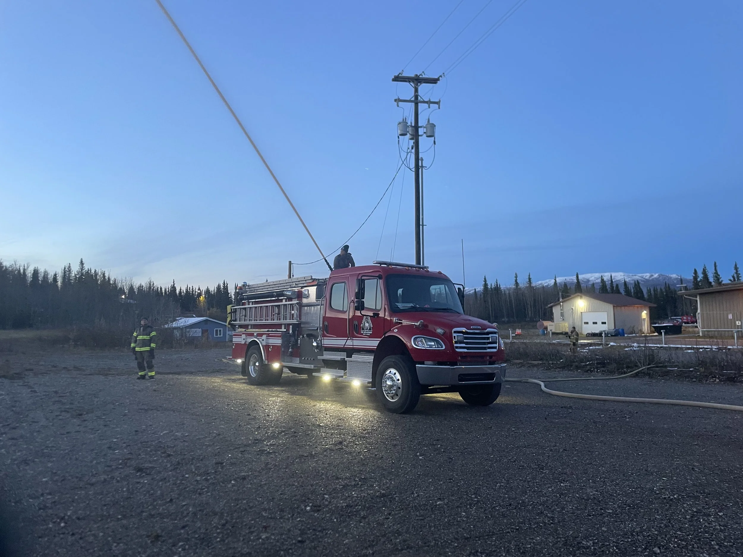 A red fire truck equipped with a ladder and emergency lights parked on a gravel surface with firefighters nearby. One firefighter is standing on the ground, and another is on top of the truck adjusting equipment. There are residential buildings and a forested area with snow-capped mountains in the background, and the sky is clear with some blue and orange hues indicating early morning or late evening.