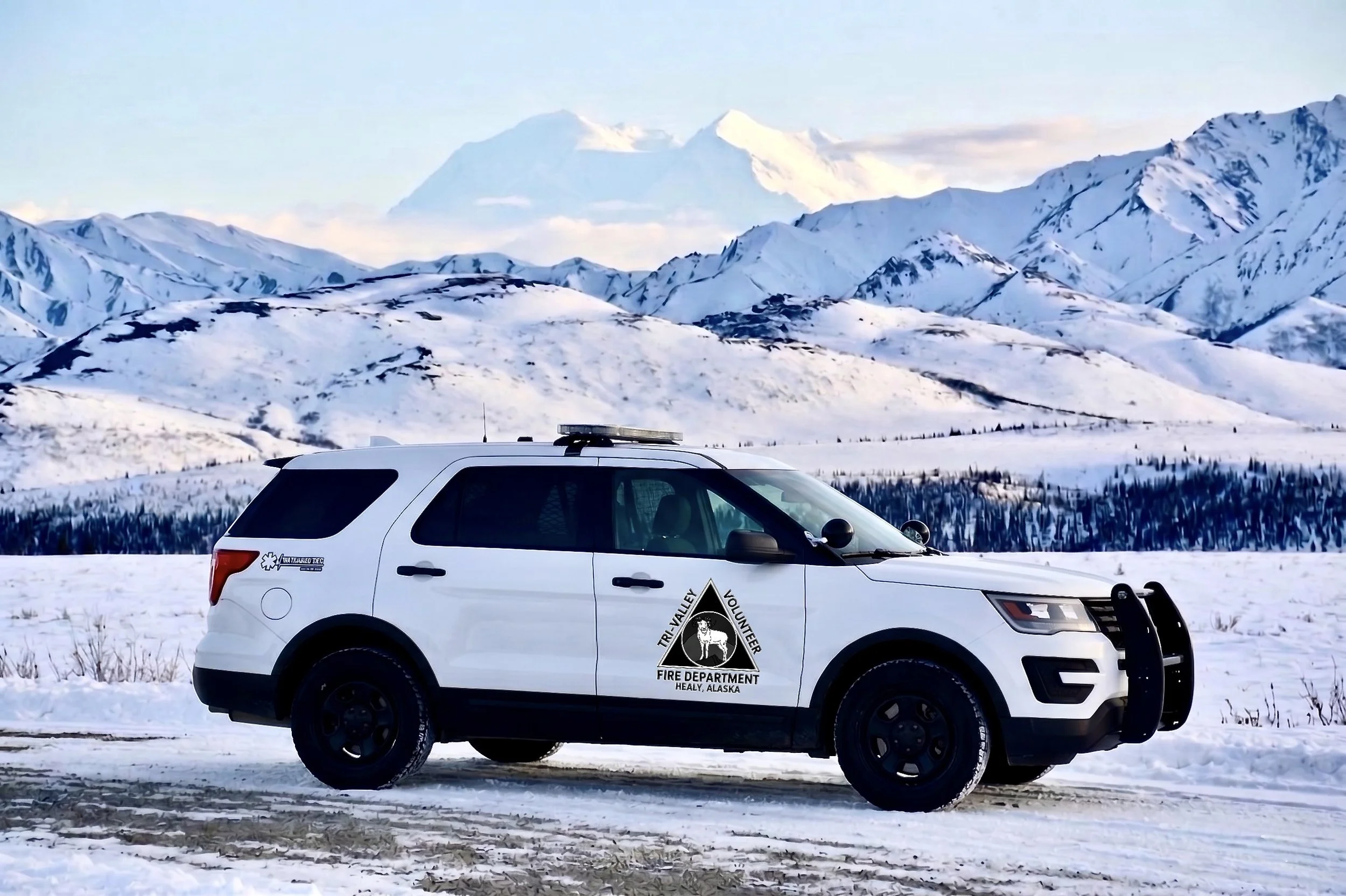 White vehicle with Tri-Valley Volunteer Fire Department emblem parked on snow-covered terrain, with snow-capped mountains and blue sky in the background.