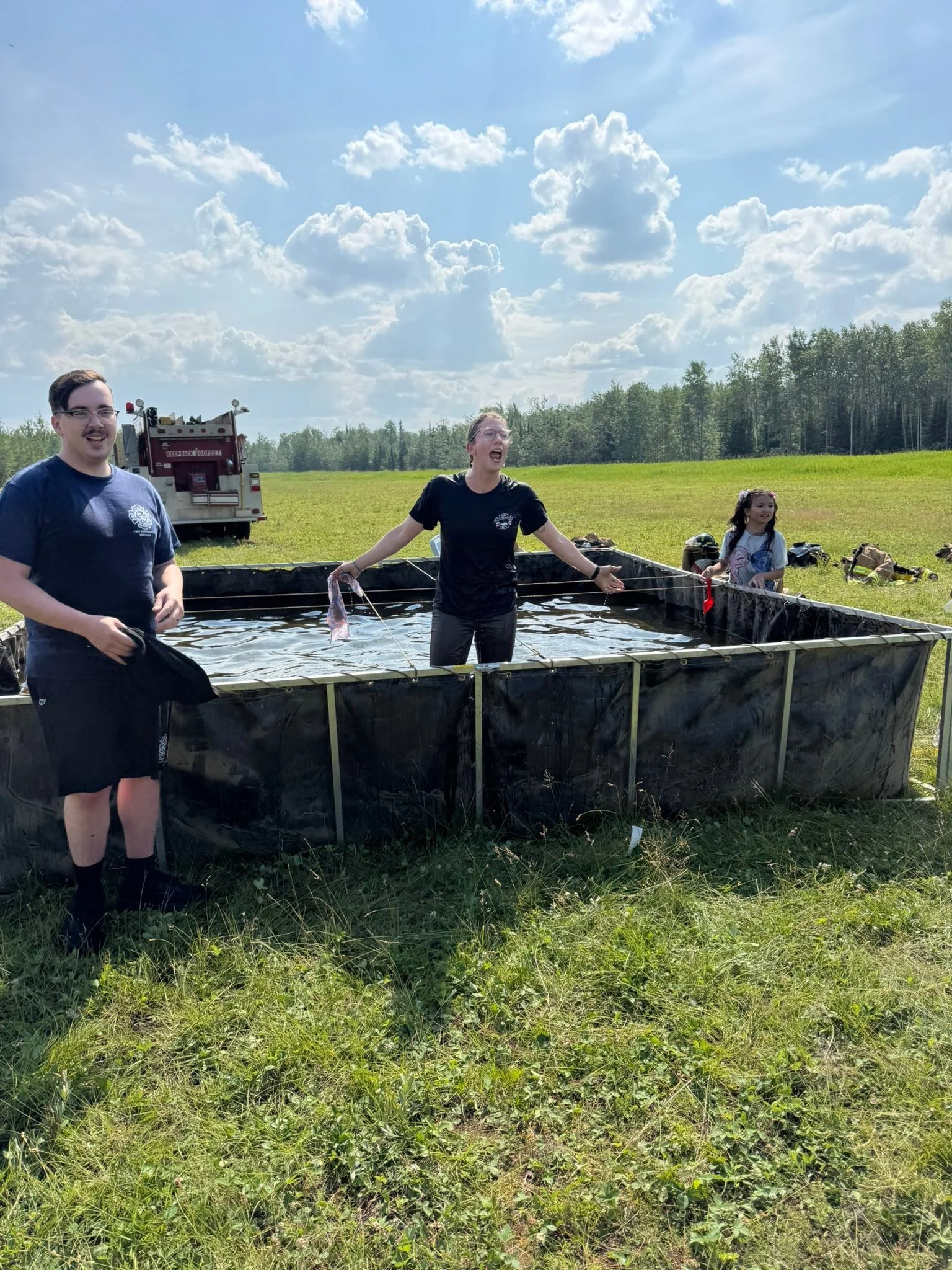 Three people, two young men and a young woman, outdoors in a grassy field with trees and a truck in the background, standing and playing in a large water tank under a sunny sky with clouds.