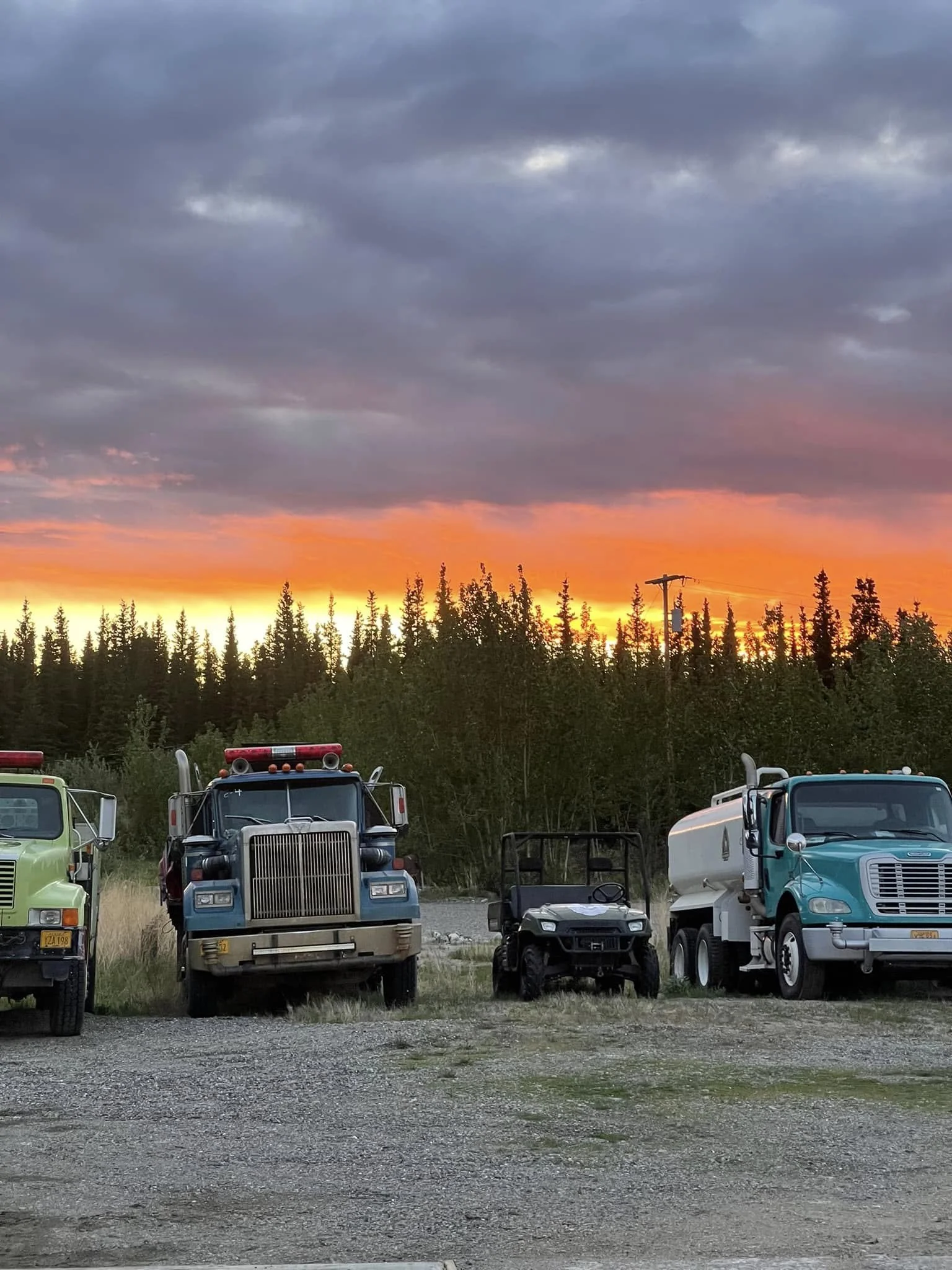 Row of trucks parked in front of a line of trees at sunset, with a cloudy sky overhead.