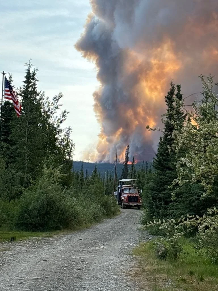 A gravel road in a forest with trees and bushes on both sides. An American flag is visible on the left. In the background, there is a large plume of smoke and fire from a wildfire, with flames visible at the base of the smoke.