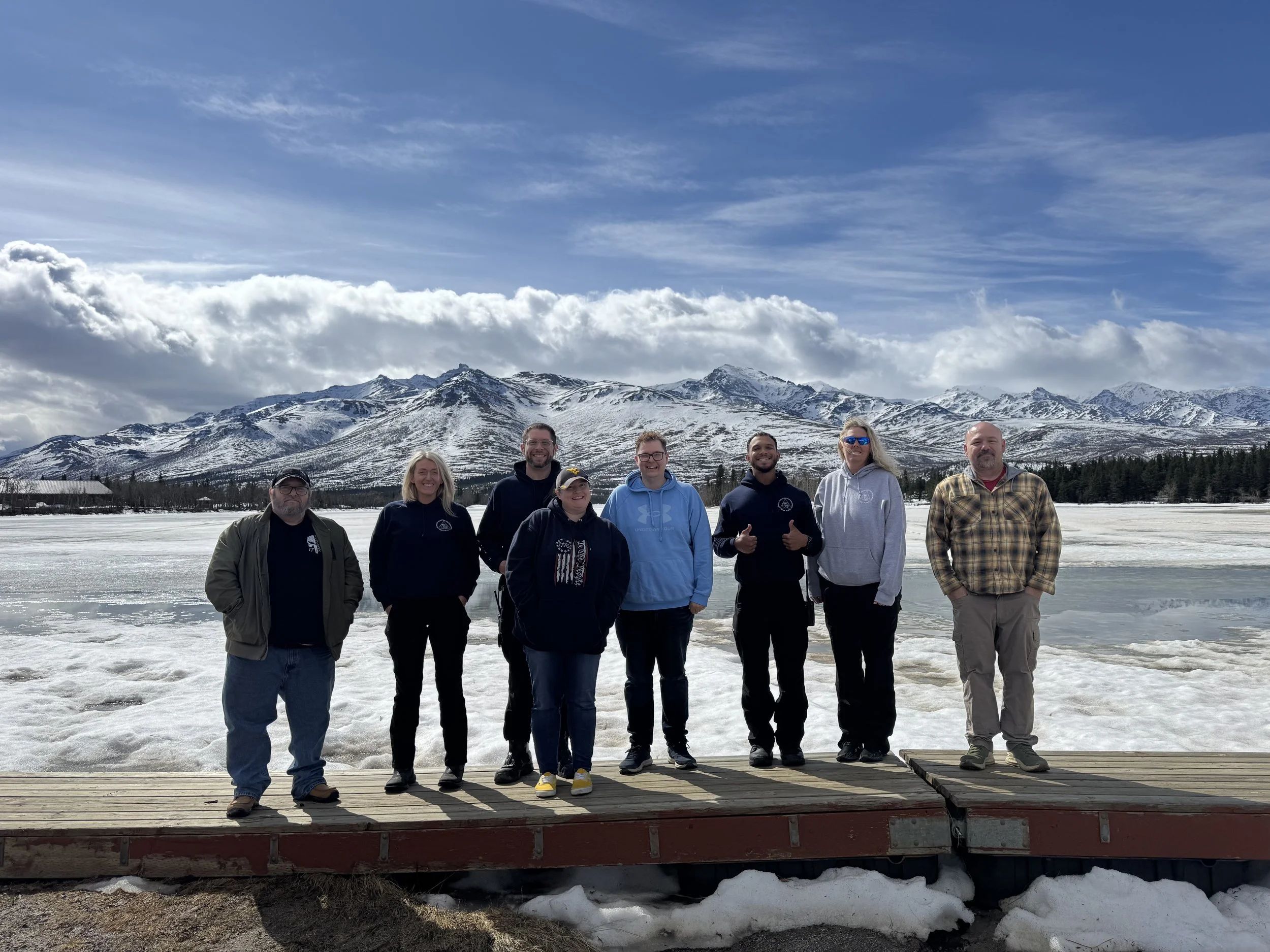 Group of nine people standing on a wooden dock with snowy mountains and a partly cloudy sky in the background.