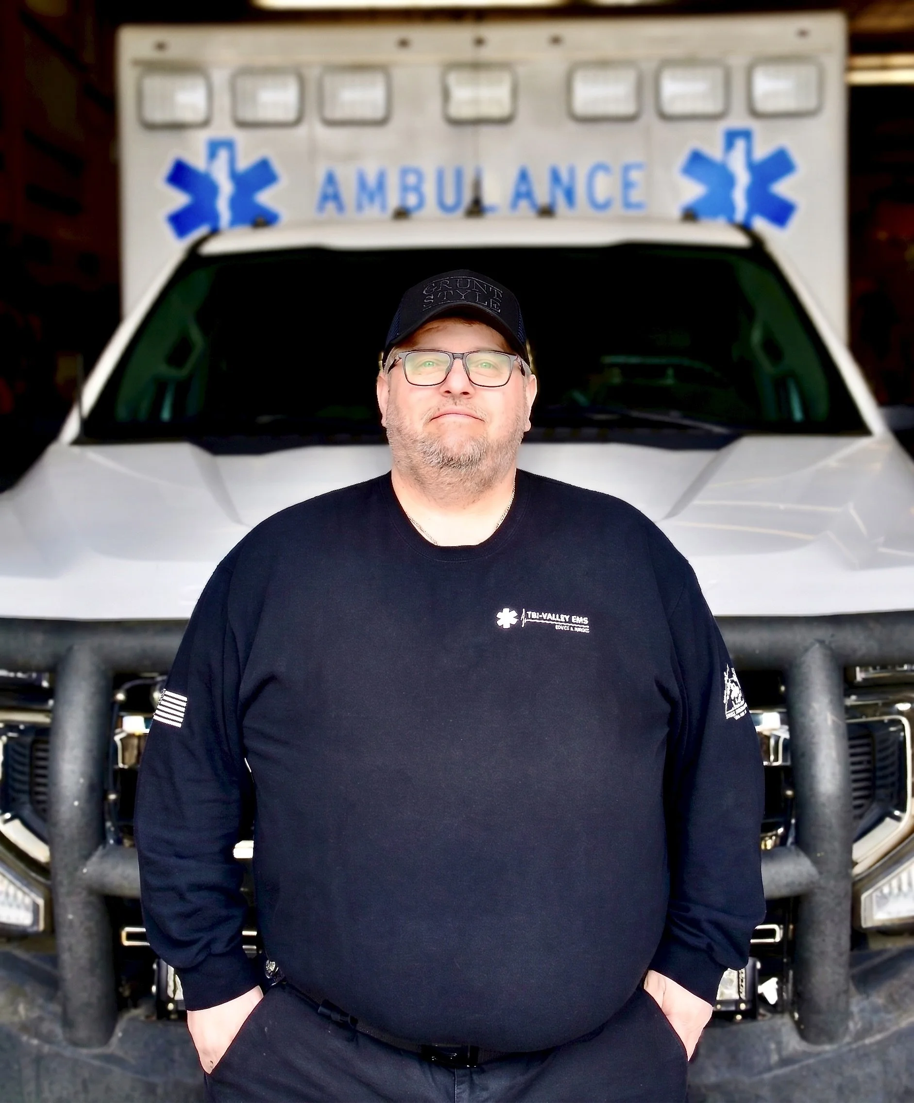 Cartoon of a smiling male emergency medical worker with a beard, wearing a white uniform and cap with medical symbols, holding a red medical kit.