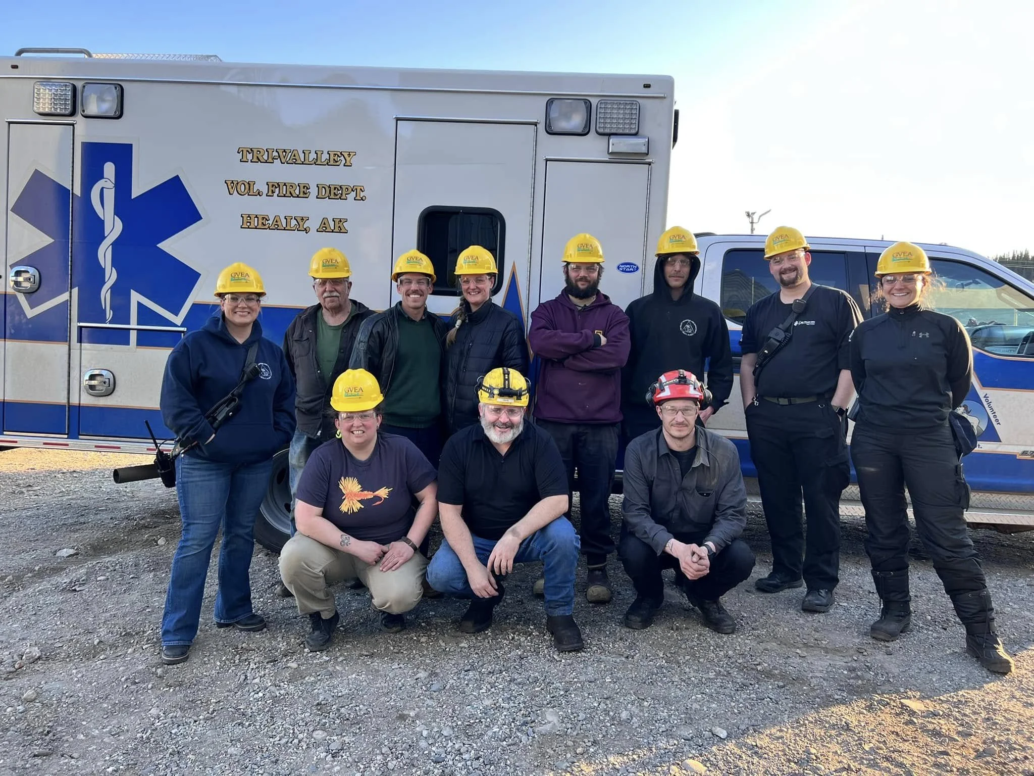 A group of ten people, some wearing yellow helmets with GVEA logo, standing in front of an ambulance marked for volunteer firefighters from Healy, Alaska. They are outdoors on gravel, with clear blue sky.