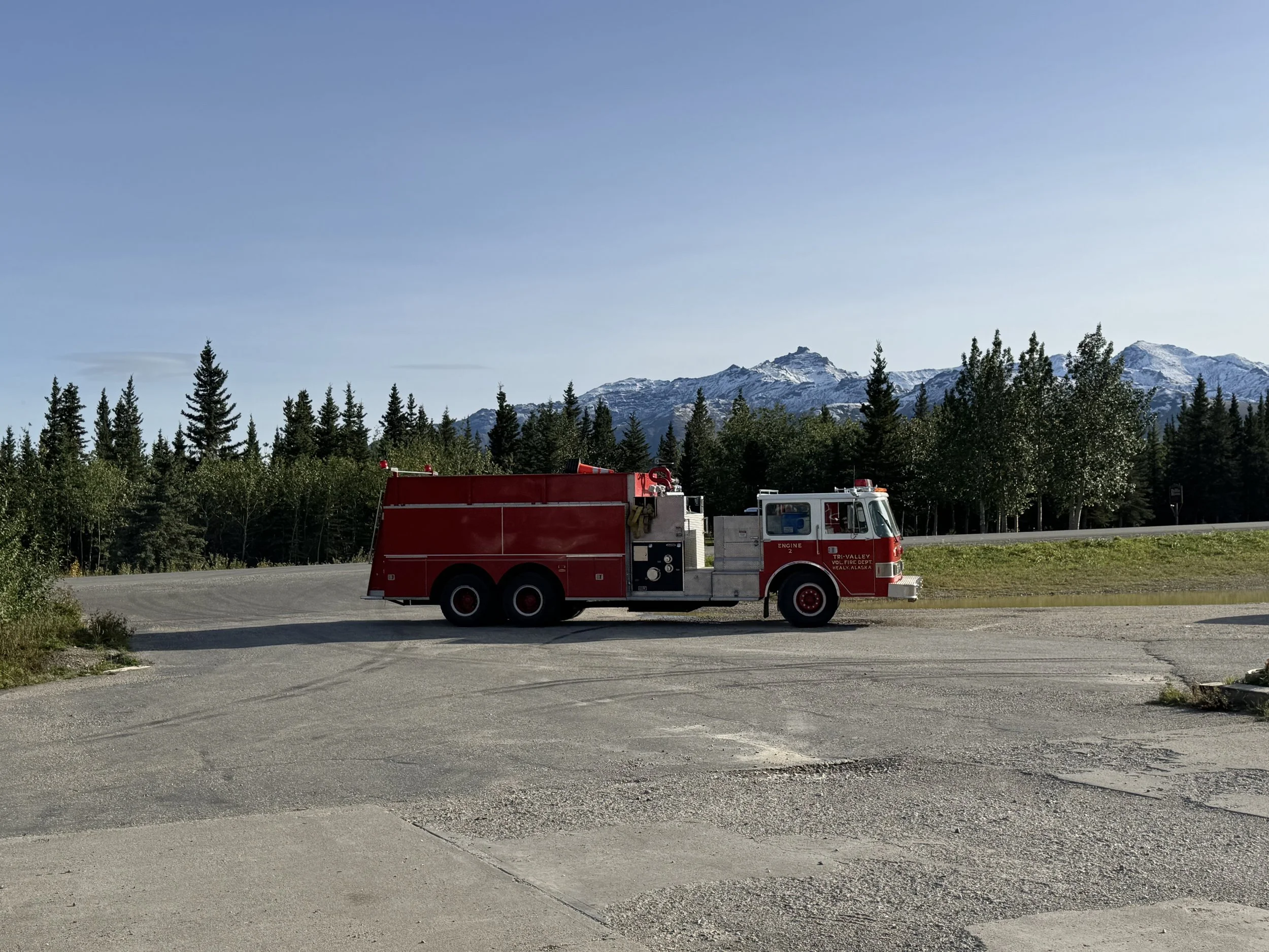 A red fire truck parked on a paved area in front of a dense forest with tall pine trees and snow-capped mountains in the background under a clear blue sky.