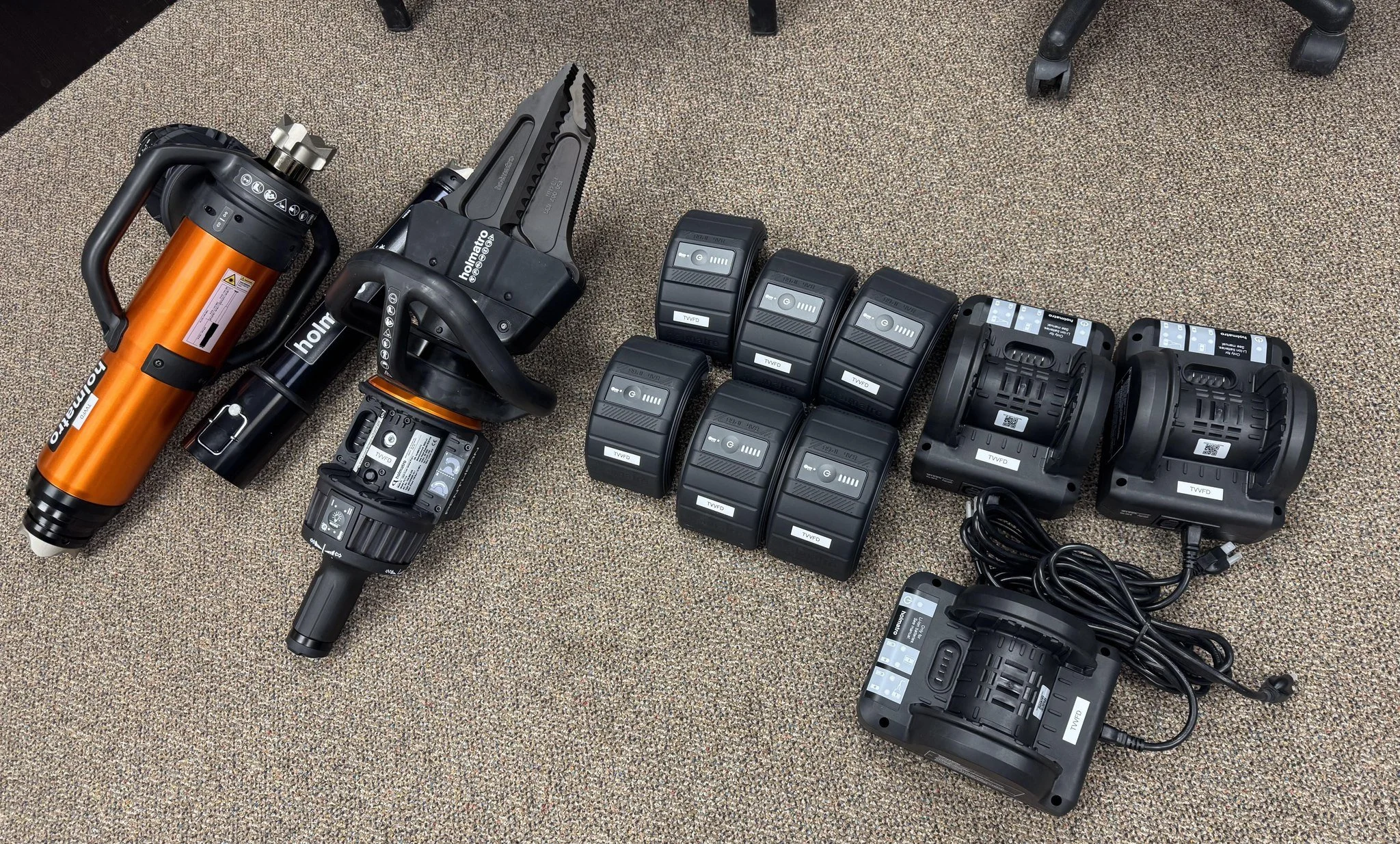Collection of camera and lighting equipment, including a large orange paint spray gun, mounted lights, and battery packs, placed on a beige carpeted floor.