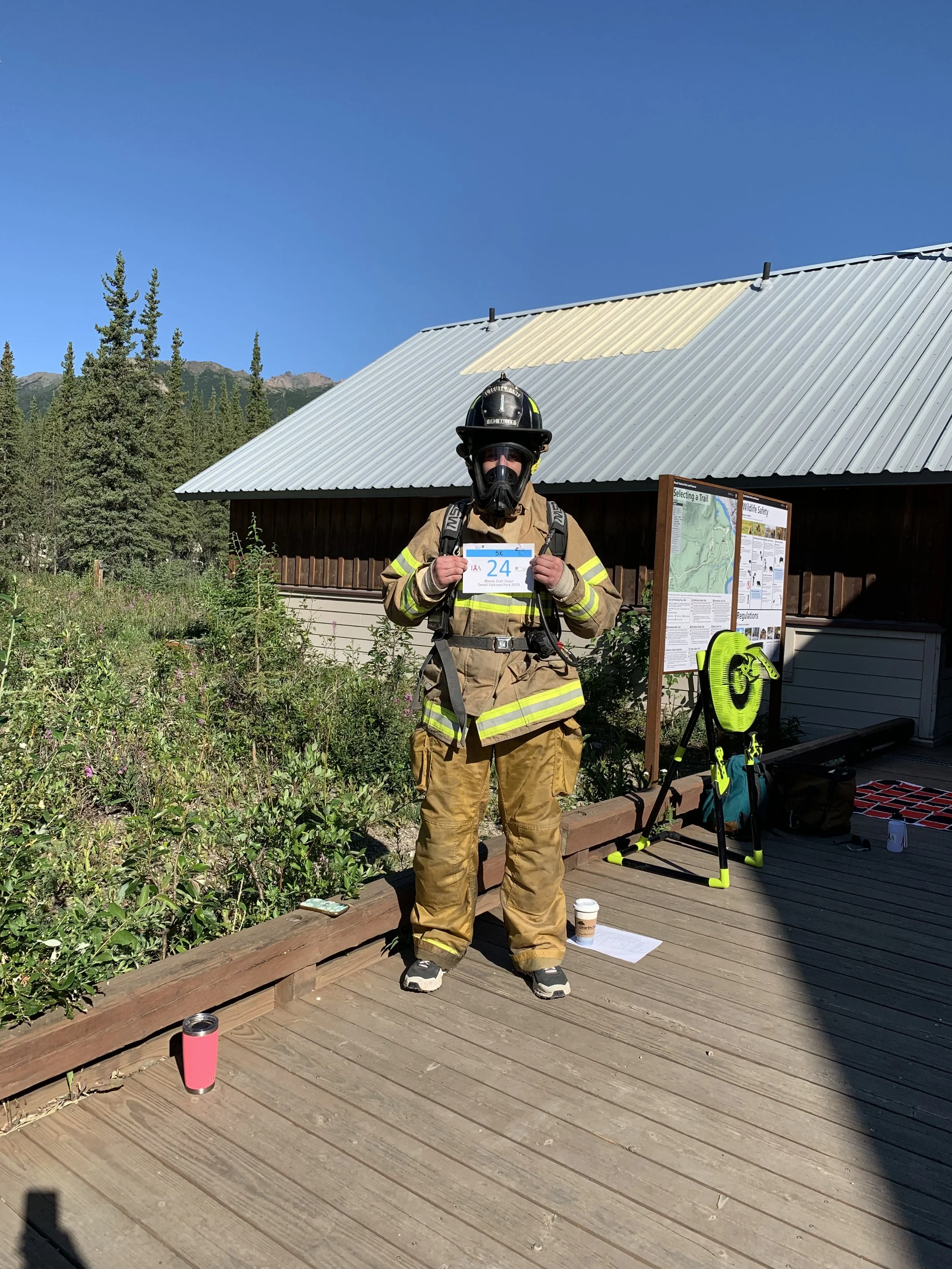 A firefighter in tan uniform and helmet standing outdoors on a wooden deck, holding a numbered bib, with a building, informational signs, and a mountain landscape in the background, under a clear blue sky.
