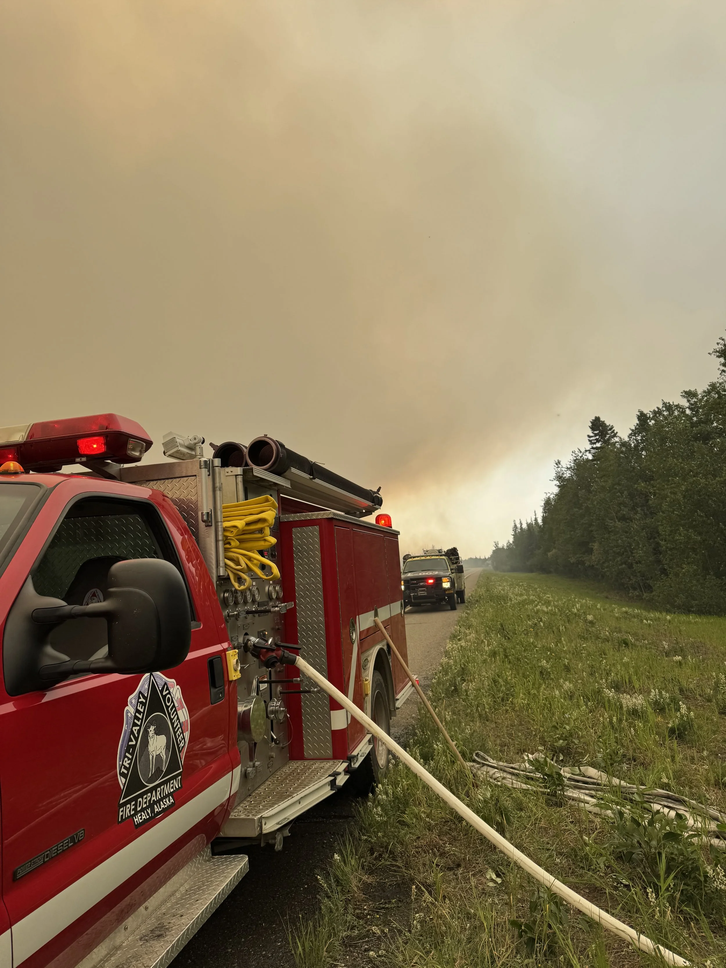 Red fire truck with firefighting equipment parked on the side of a rural road with smoke-filled sky and trees nearby.